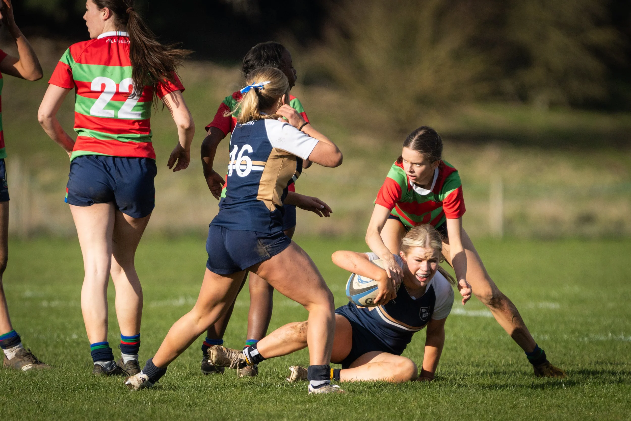 Women playing rugby on a grassy field, some are tackling and others are running with the ball, wearing sports uniforms in red, green, blue, and beige.