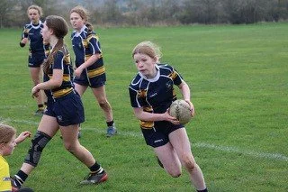 Girls playing rugby on a grassy field, one girl runs with the ball while others chase and support.