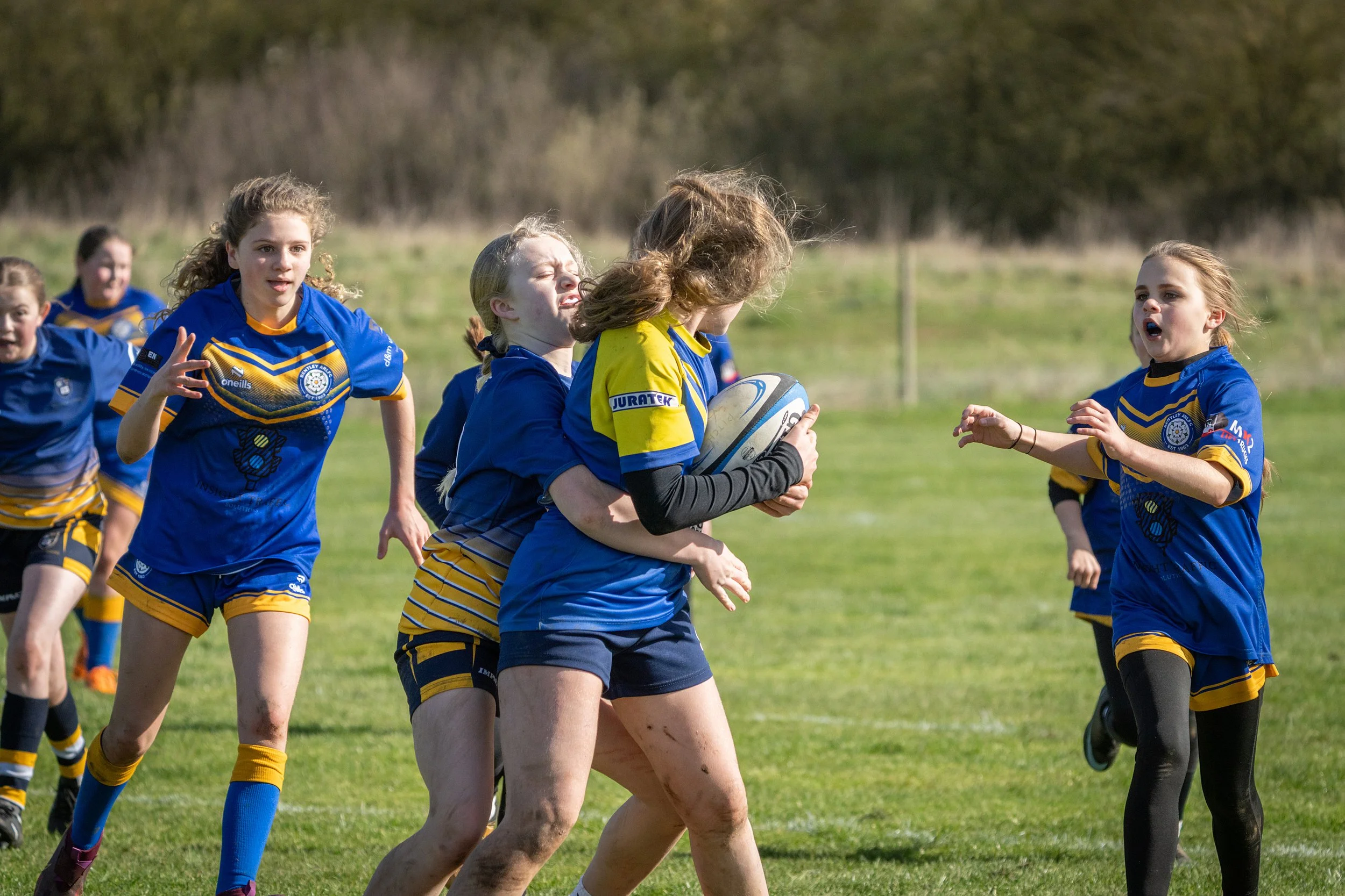 Young girls playing rugby outdoors on a grassy field, wearing blue and yellow uniforms, with one girl holding a rugby ball.