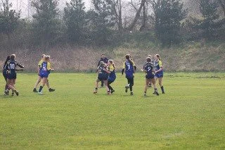 Rugby players in a match, with one player holding the ball and others tackling or defending, on a grassy field.