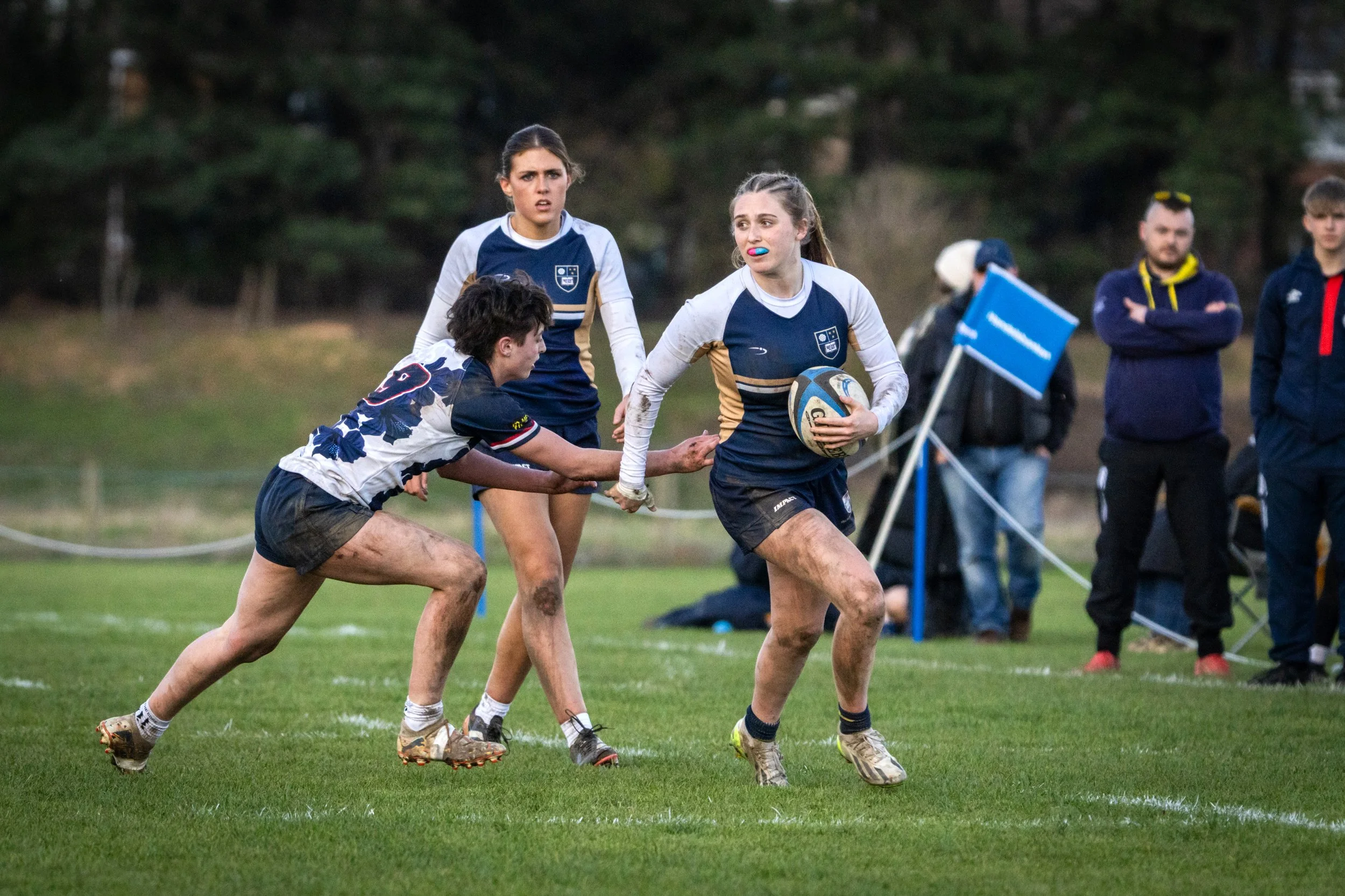 Two female rugby players, one with a ball, are running on a grassy field during a match. A person in a white and navy jersey is tackling a player in a navy and white jersey. In the background, there are spectators and trees.