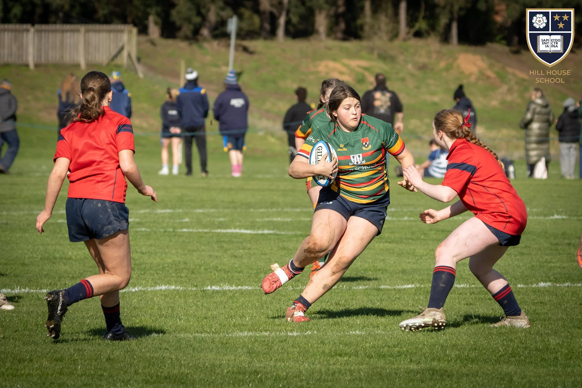 Young female rugby players in green and red jerseys competing on a grassy field, with onlookers and trees in the background, and a school crest in the upper right corner.