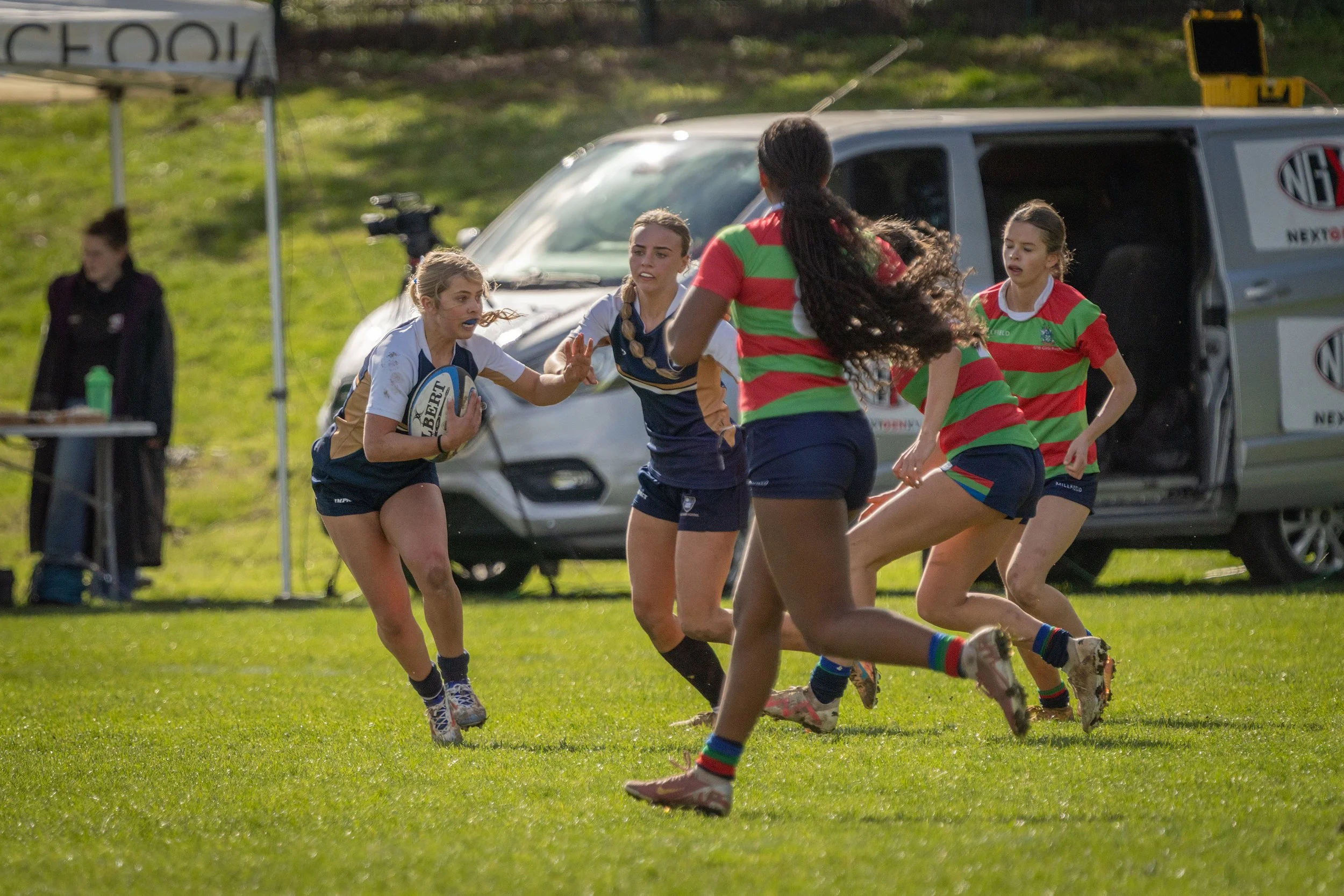 Women playing rugby on a grassy field with a van and spectators in the background.