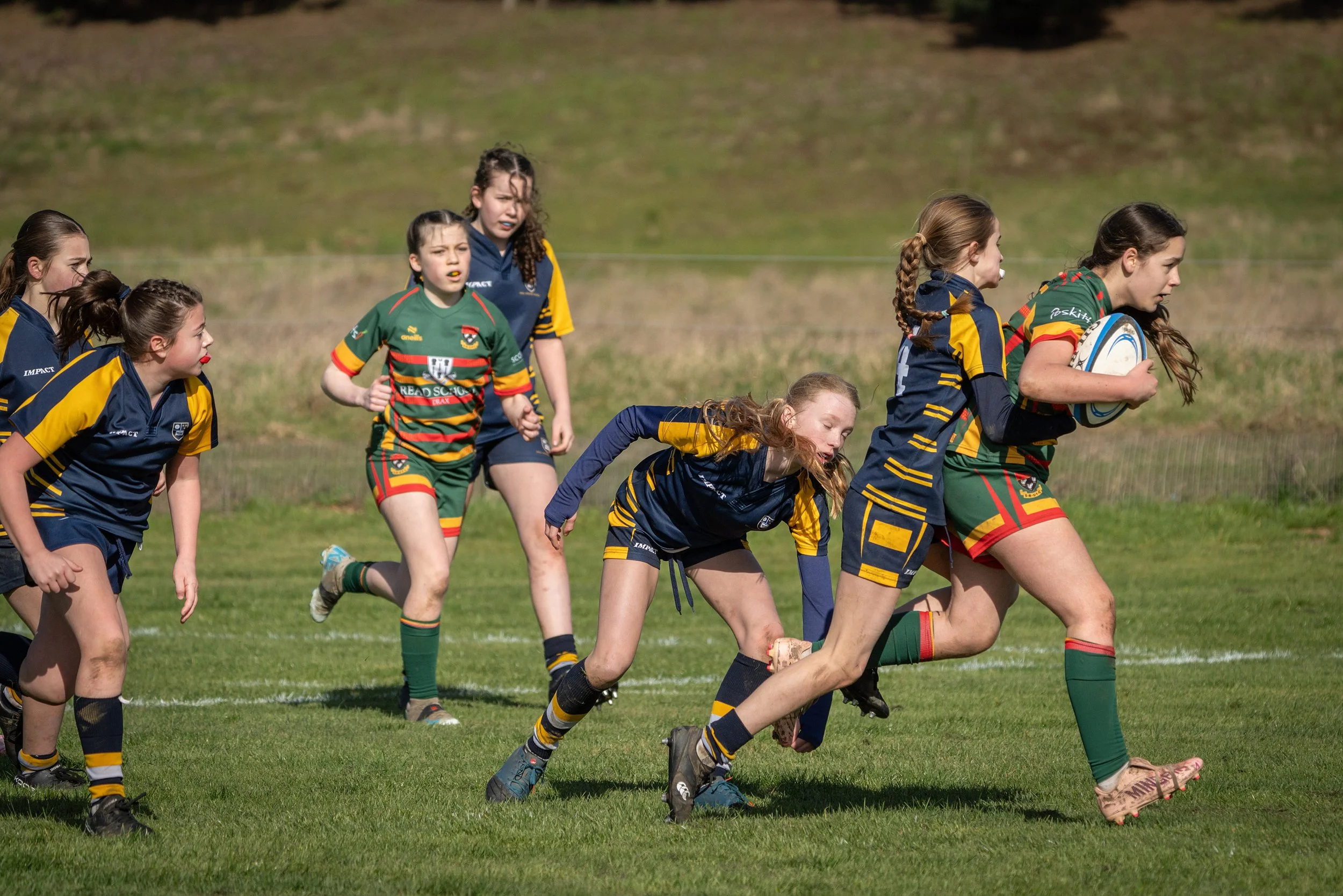 Young girls playing rugby on a grassy field, some wearing green and others wearing navy and yellow jerseys.