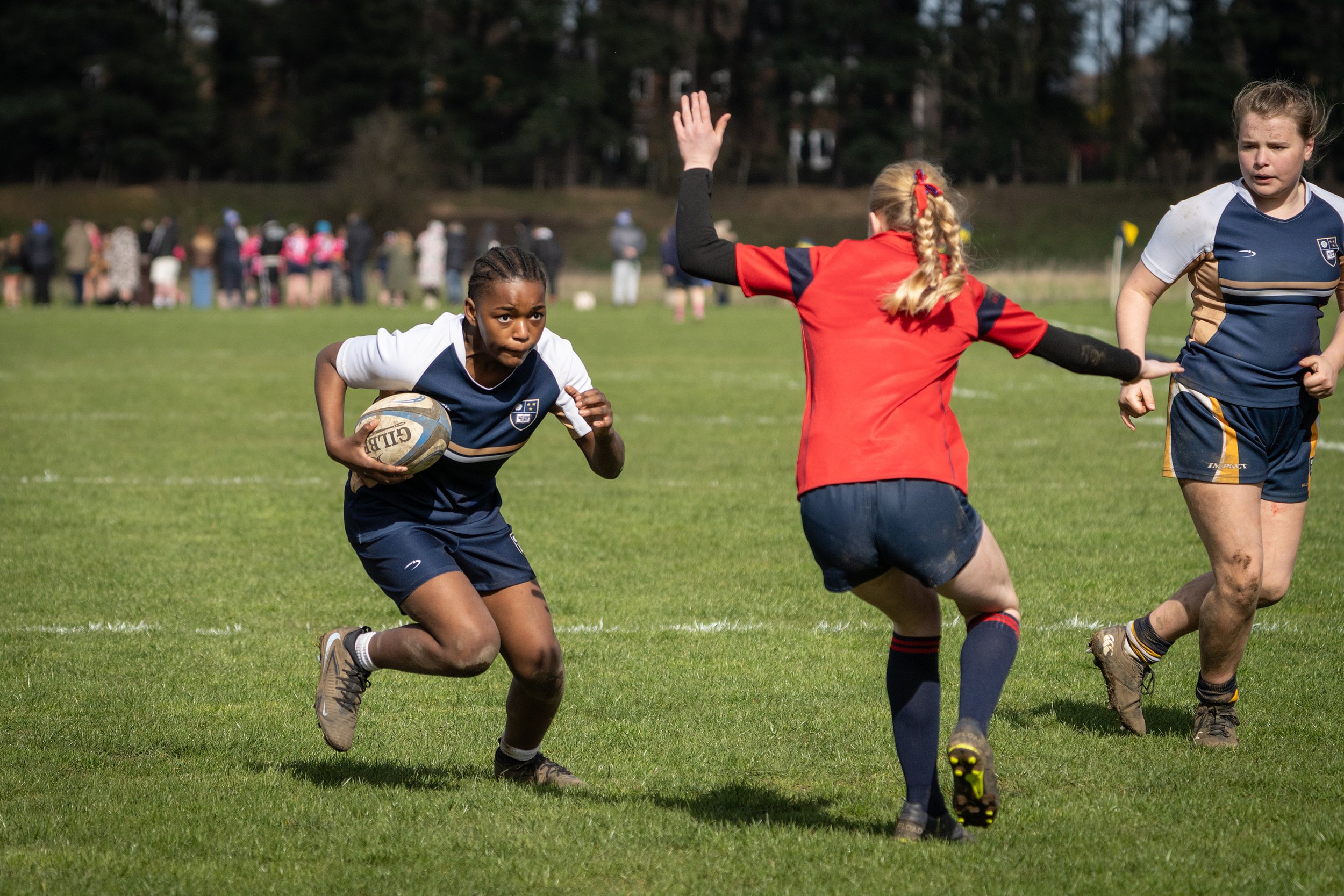 A girls' rugby game in progress on a grassy field with players wearing navy blue and white or red jerseys. One girl with braided hair and a determined expression holds a rugby ball, running forward. An opponent in a red jersey raises her arm for a ta