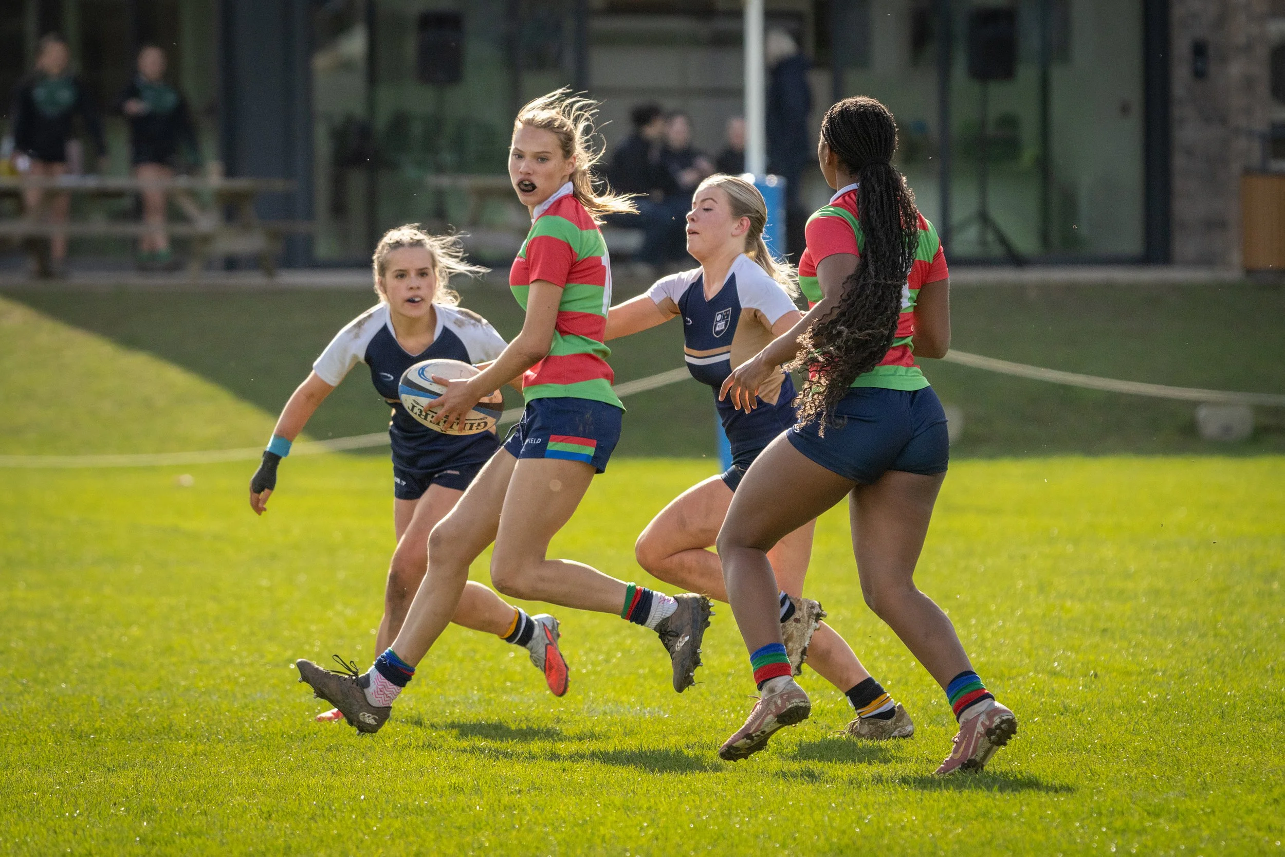 A group of young women playing rugby on a grassy field, with one holding a rugby ball and others running alongside her.