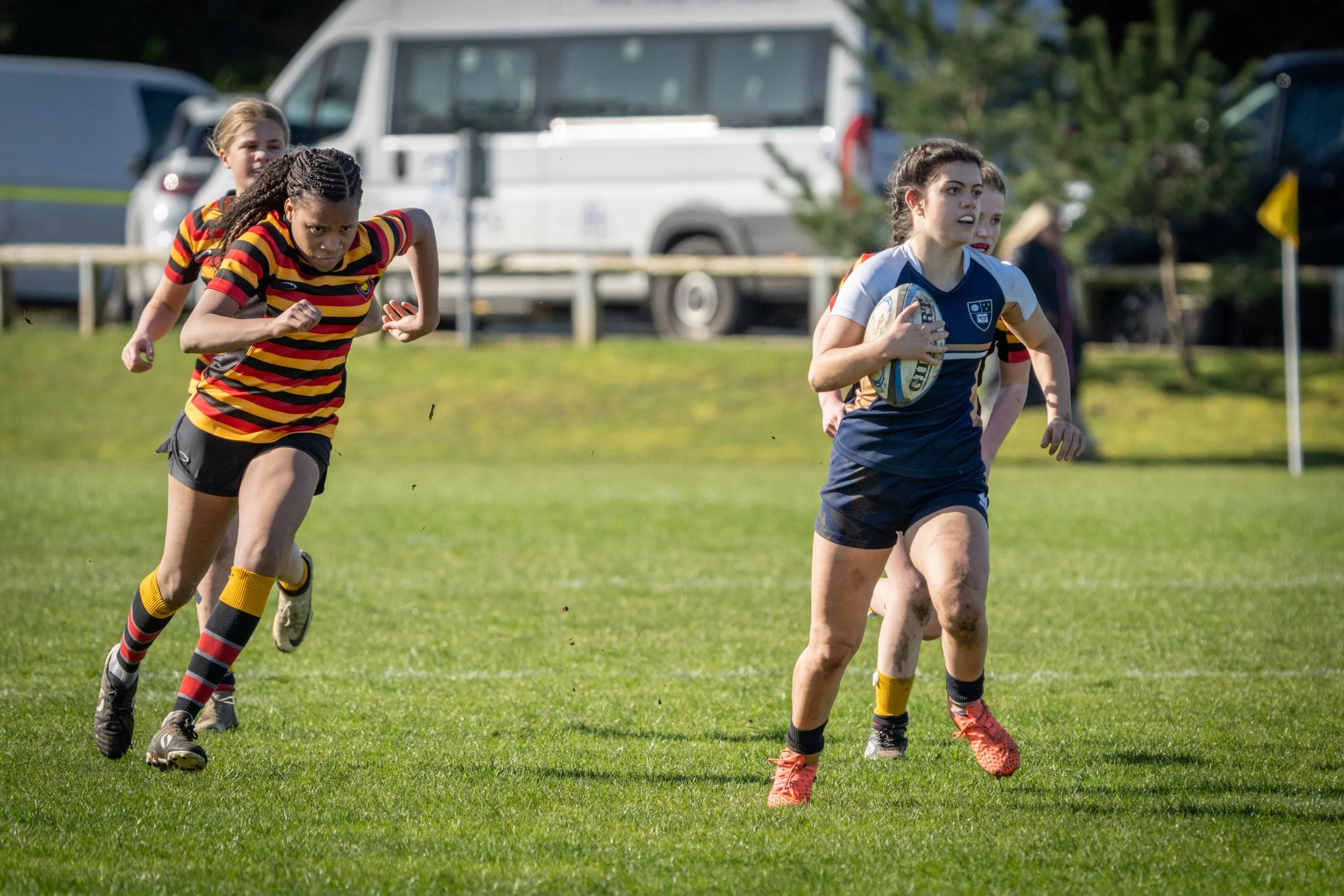 Young female rugby players competing on a grassy field, with one holding a rugby ball and others running behind.