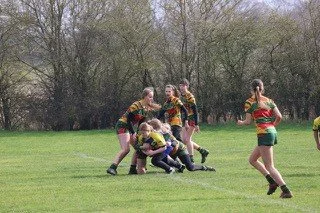 Women playing rugby on a grassy field with trees in the background.