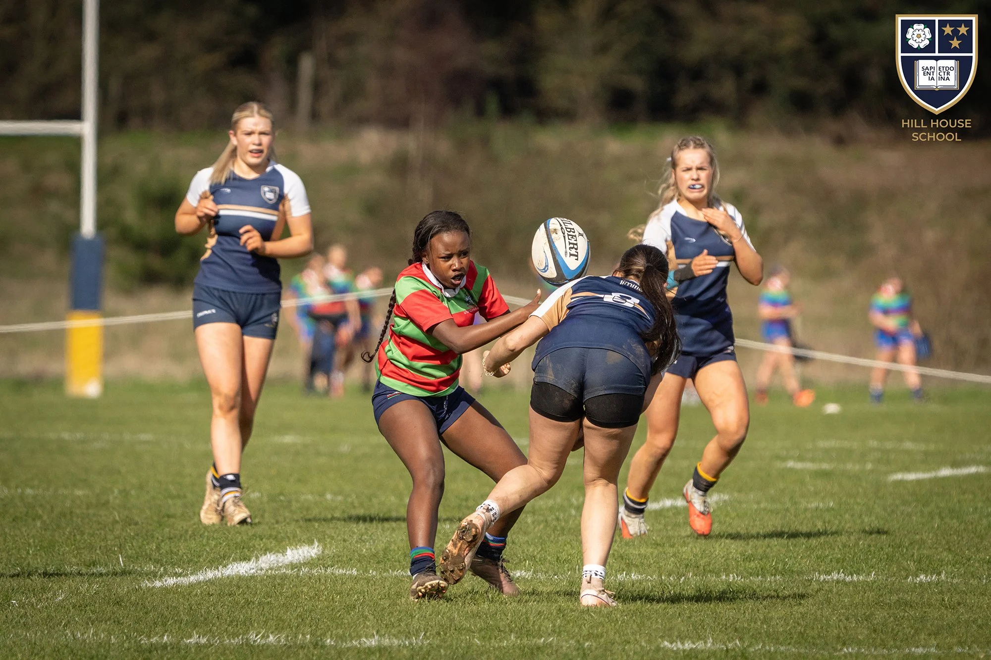 Girls playing rugby on a grassy field, with a player in a green and red jersey catching the ball while others in blue jerseys run nearby, and a blurred background with spectators and trees.