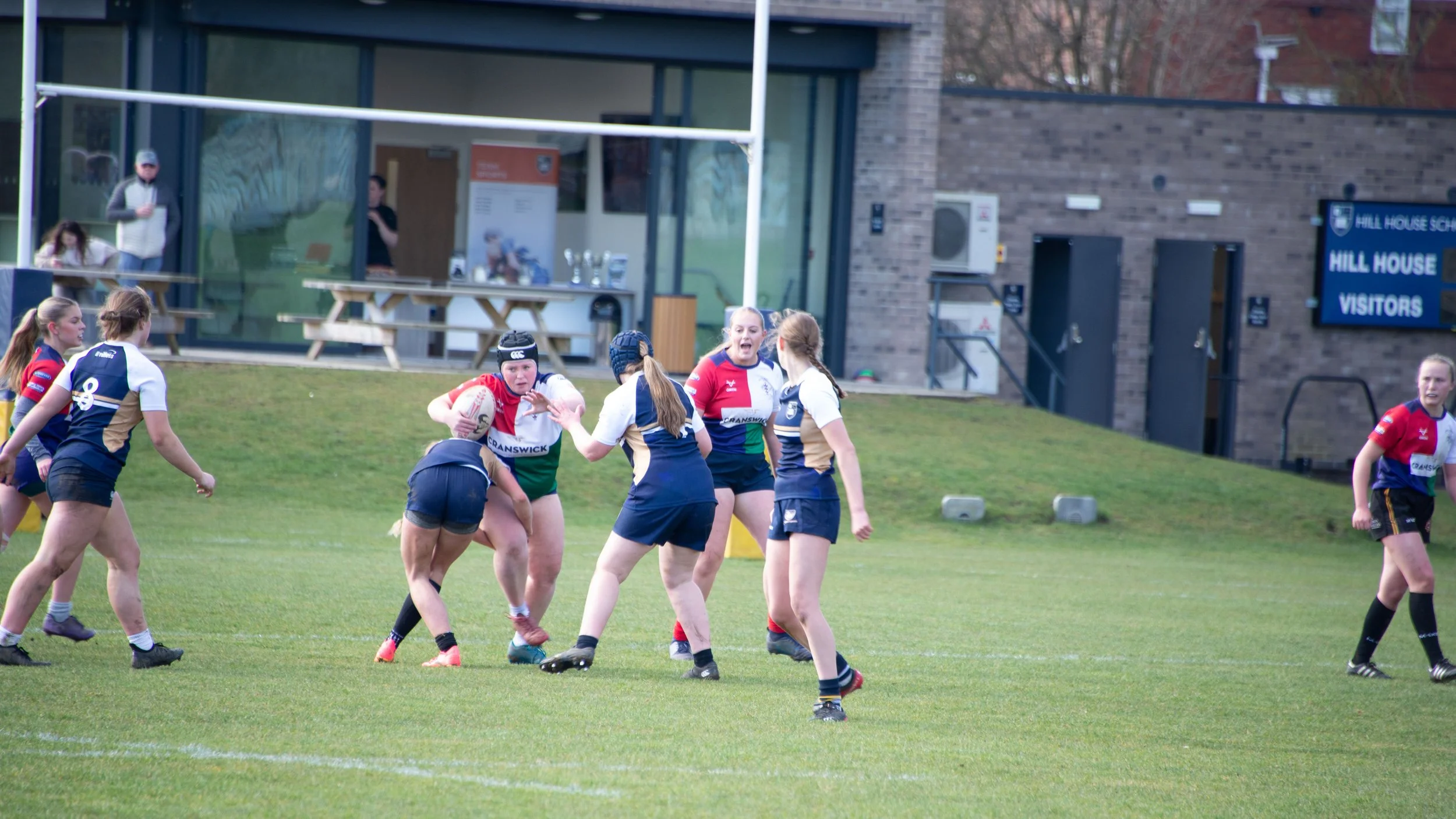 Girls rugby team playing on a grassy field outside a building with spectators watching.