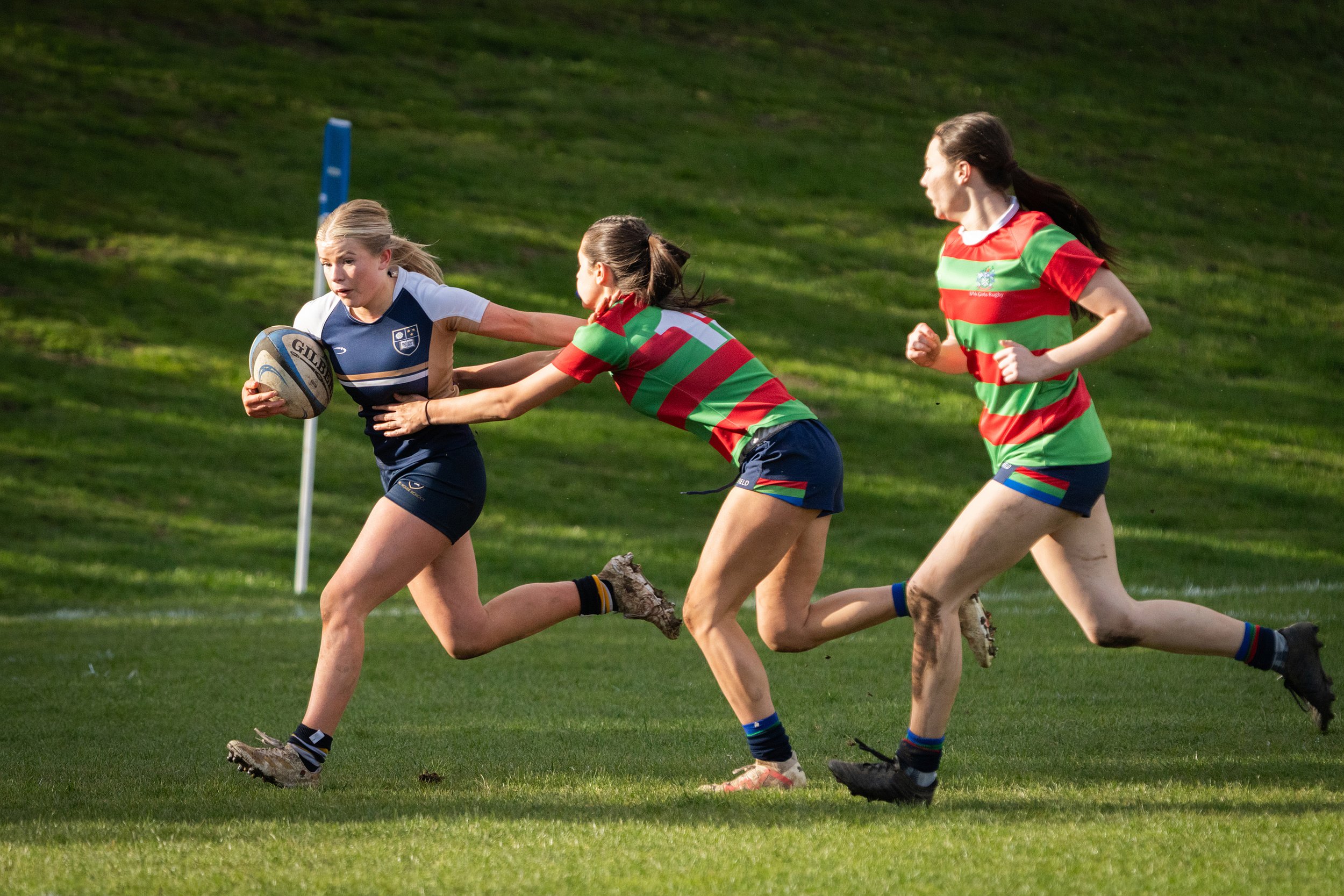 Women playing rugby, running on a grass field, with three women in focus, one holding a rugby ball, wearing sports uniforms.