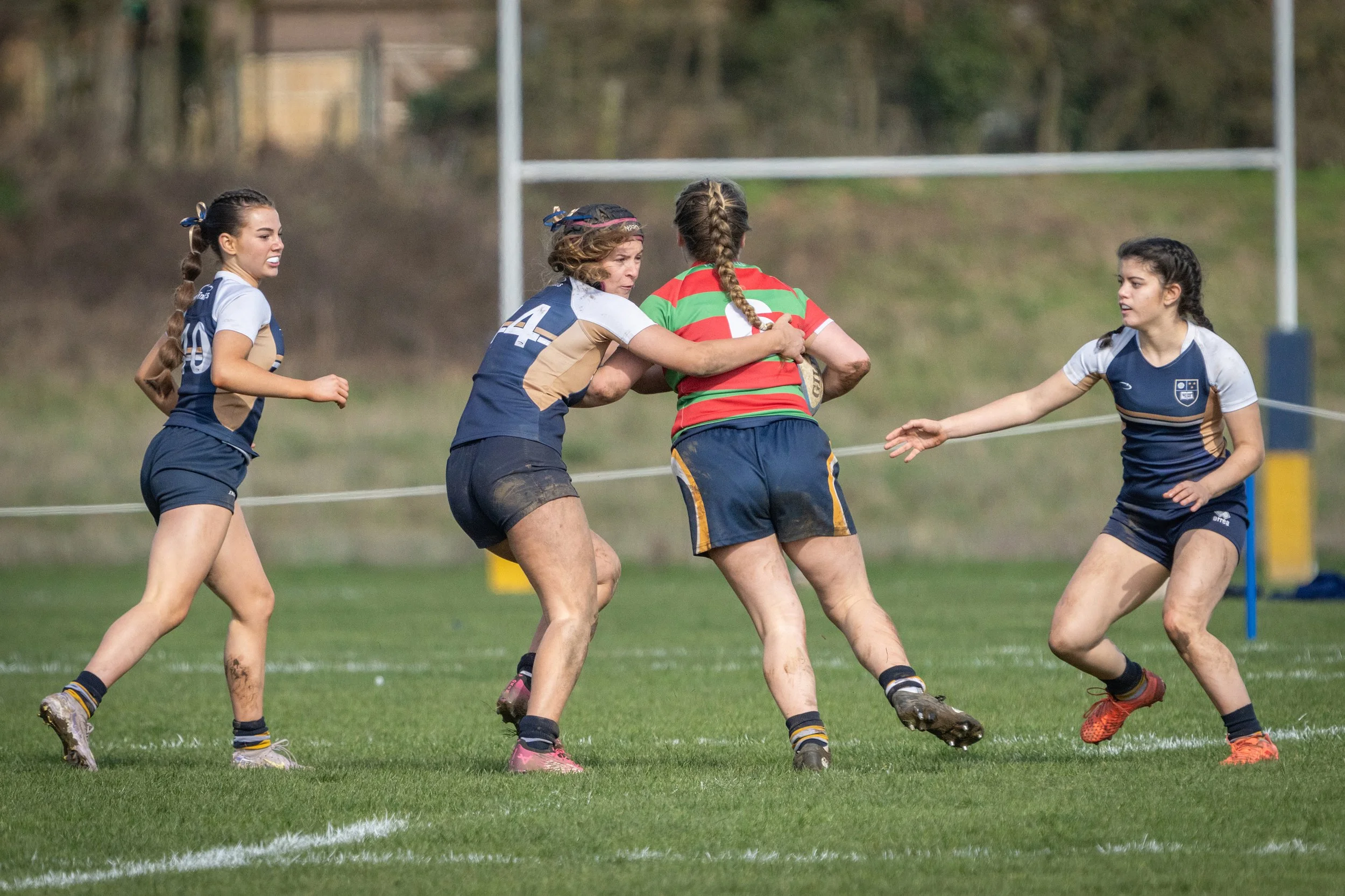 Women playing rugby on a grassy field, one woman in a red and green striped jersey holding a rugby ball, surrounded by three women in blue and beige jerseys.