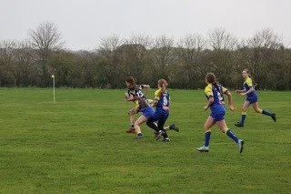 Five girls playing rugby on a grassy field under an overcast sky.