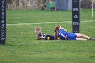 Two young rugby players lying on the grass near the goal post, one on their stomach and the other on their side, during a game.