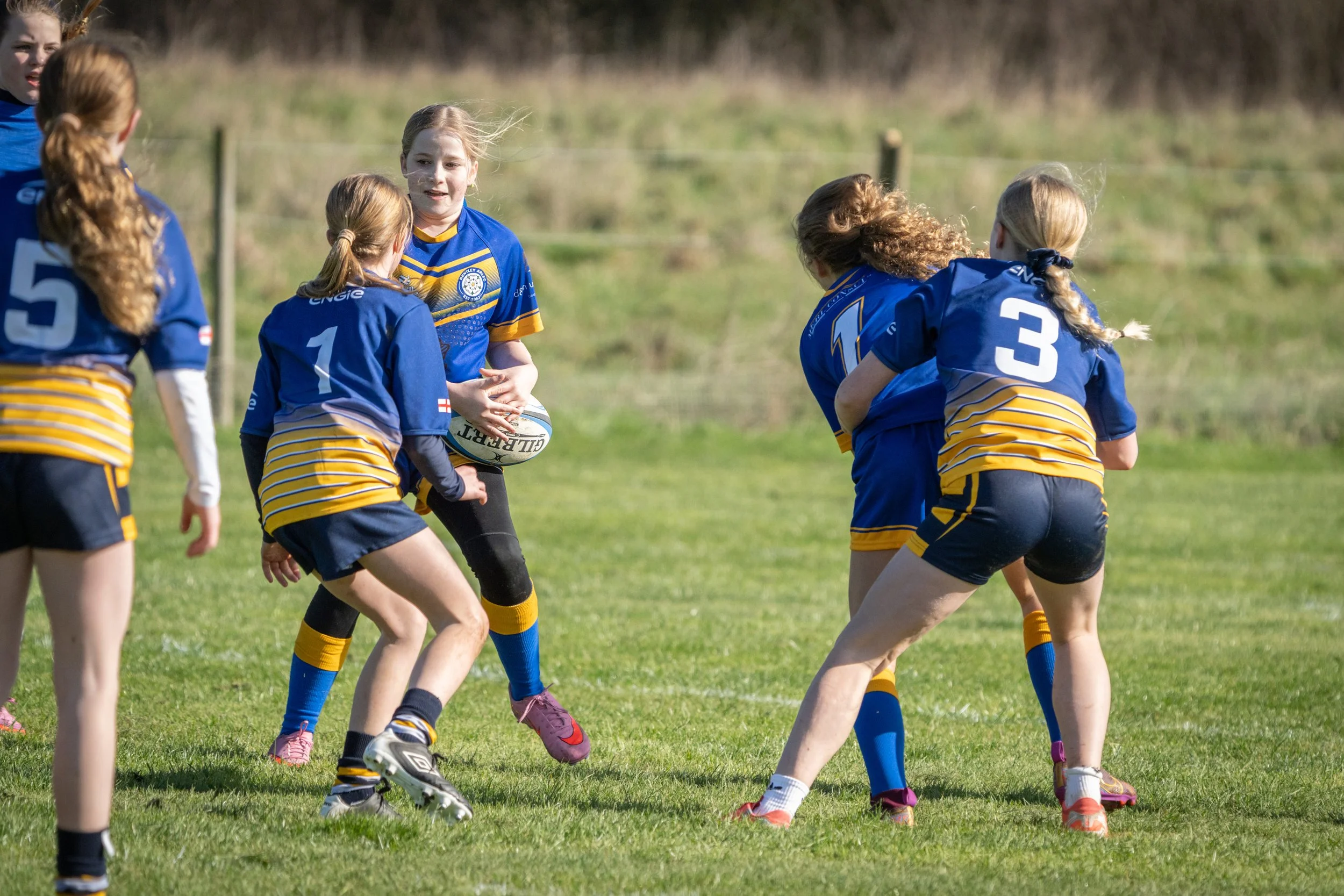 A group of young girls playing rugby on a grassy field during daytime, wearing blue and yellow uniforms.