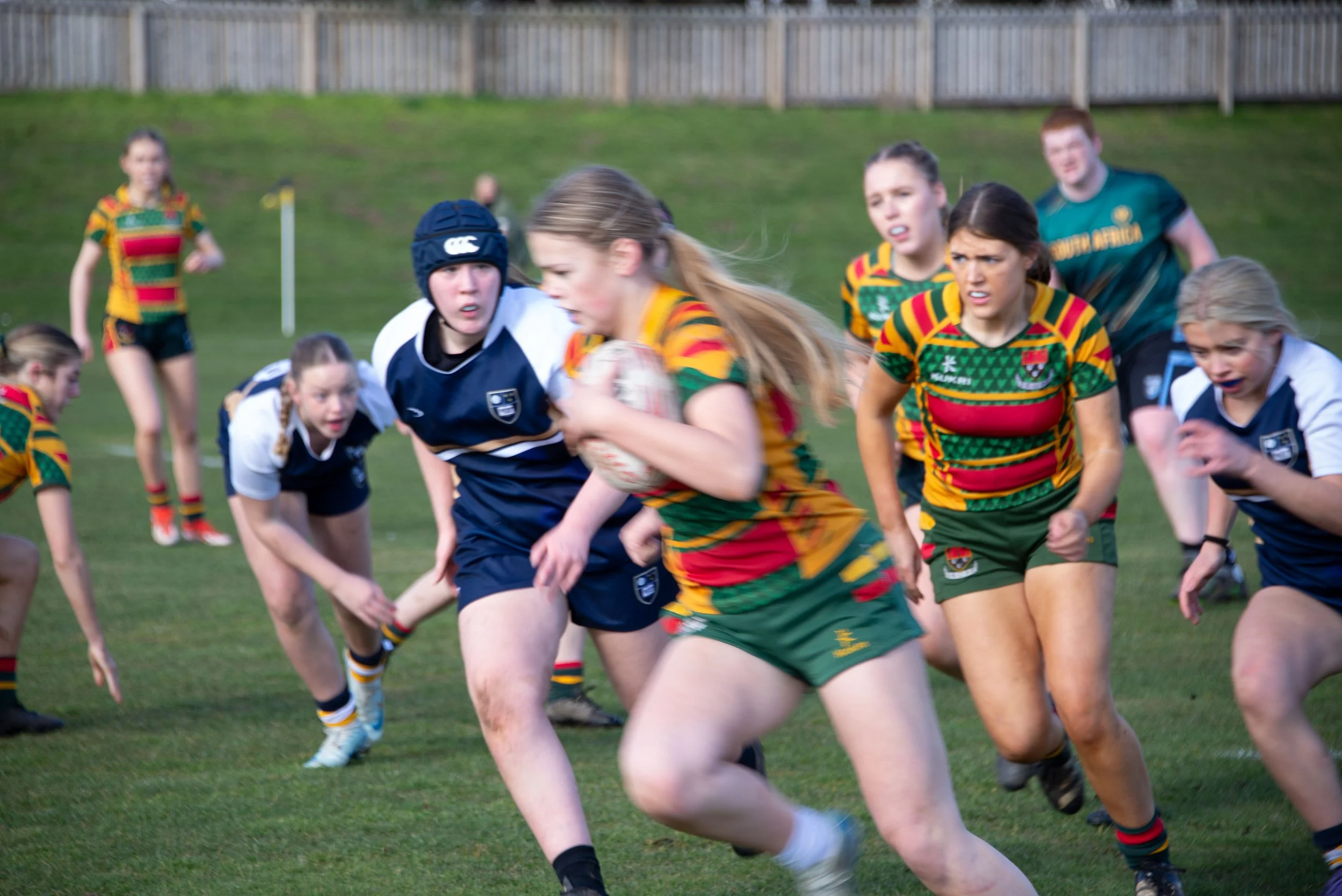 A women's rugby match with players in colorful uniforms and a female player carrying the ball across a grassy field, with other players pursuing.