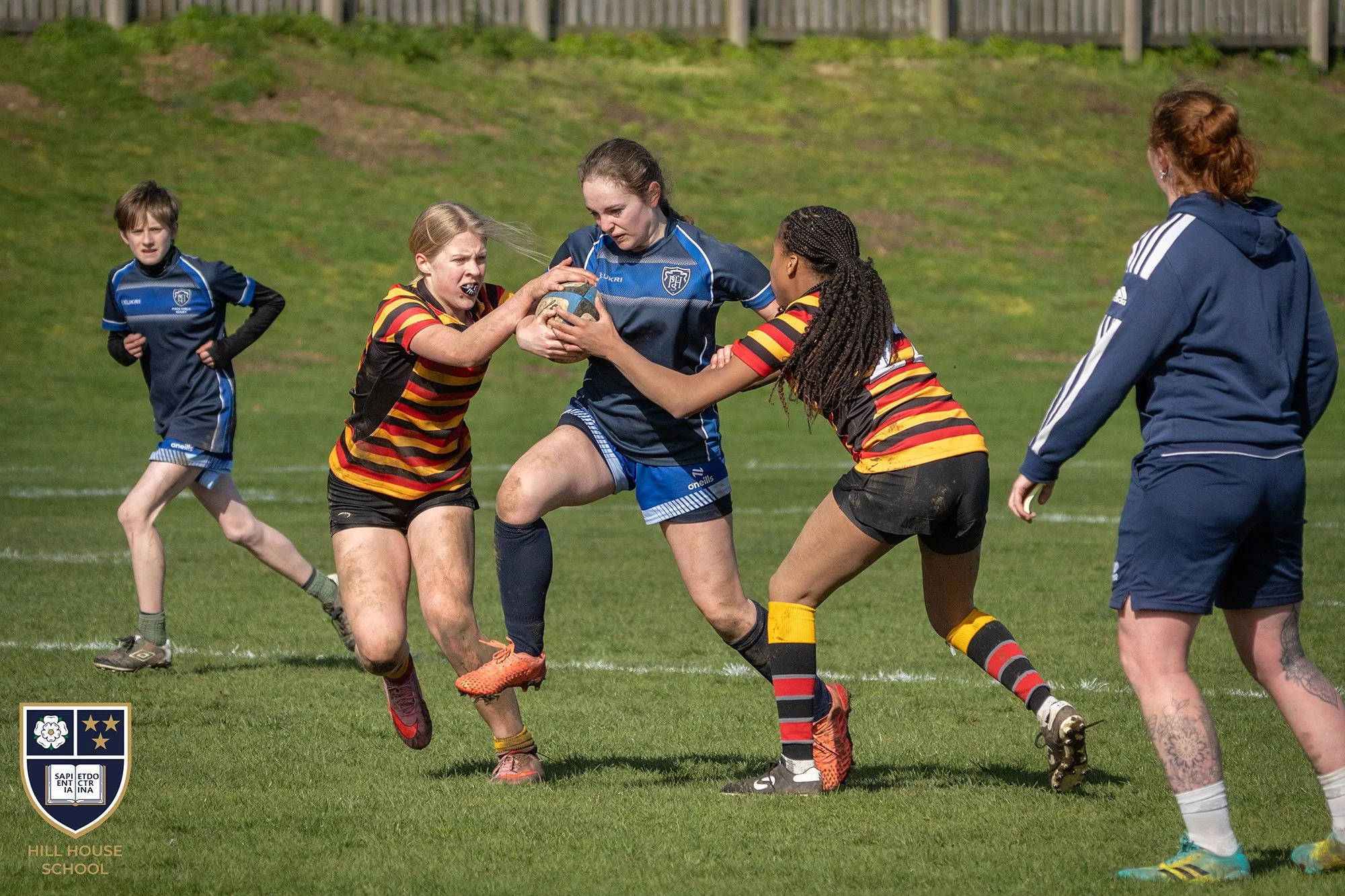 Young girls playing rugby on a grassy field, with one girl in a blue jersey holding the rugby ball while others try to wrest it away, and a girl in a striped jersey attempting to tackle her. A coach or referee stands nearby. The background features a
