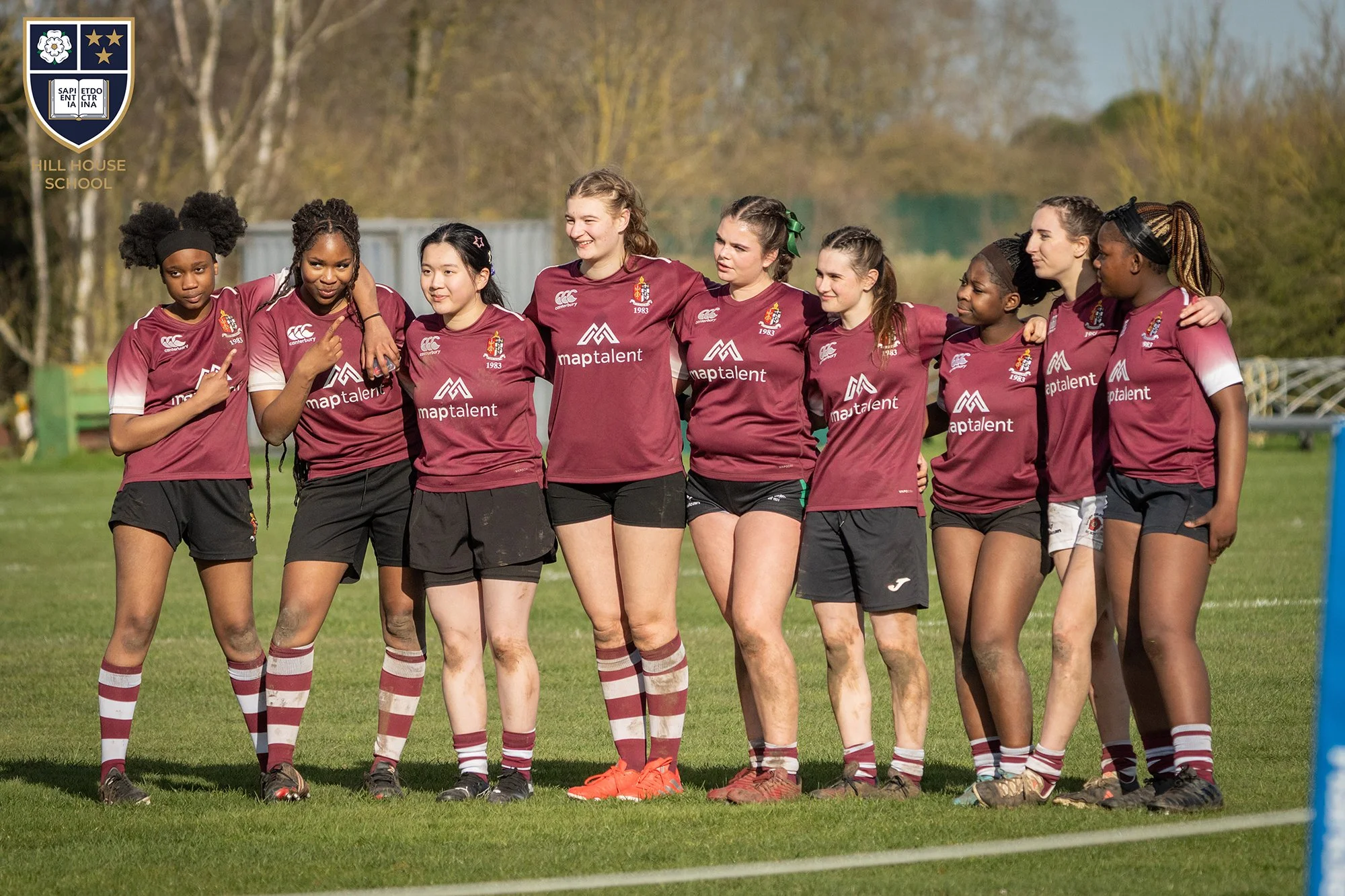 Group of young female rugby players standing on a field with arms around each other, wearing maroon jerseys and shorts, some with hands on shoulders, smiling, and looking towards the right side of the image, with trees in the background.