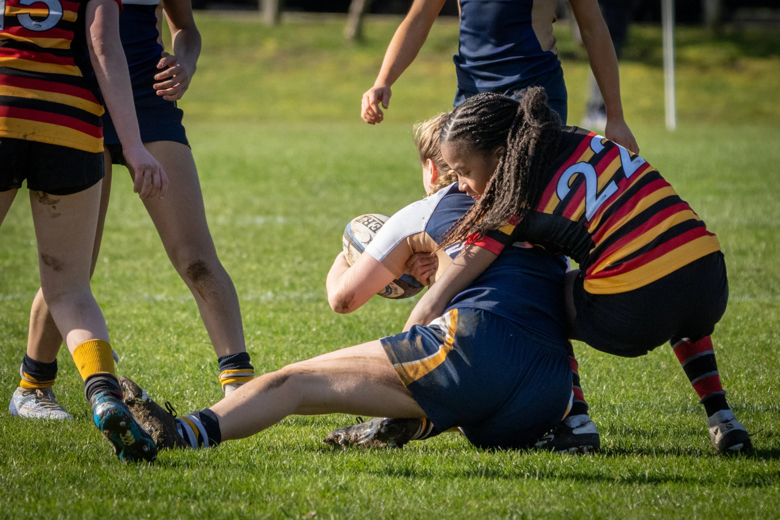 Women rugby players in a scrum during a match, with dirt on their legs, on a grassy field.