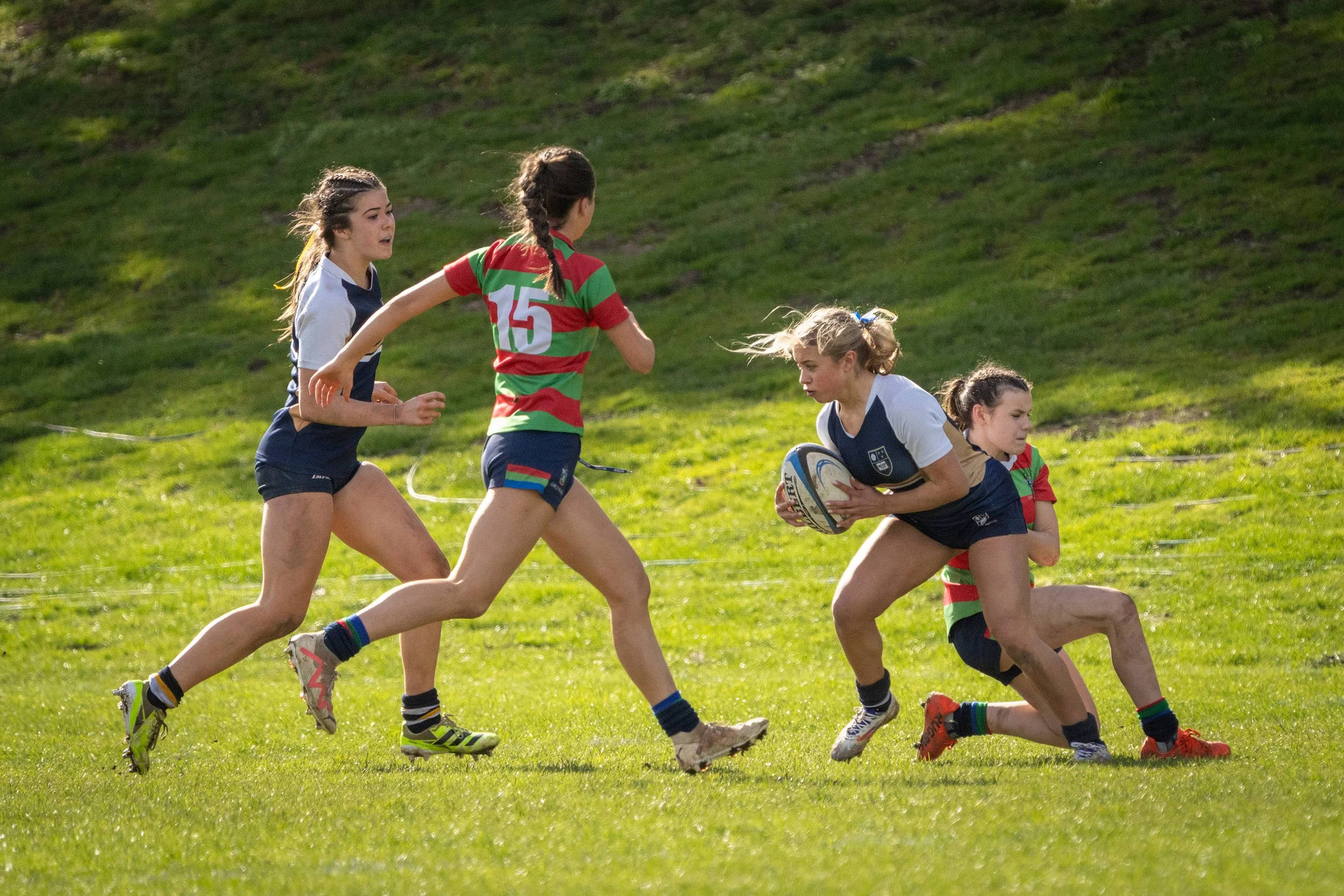 Girls playing rugby on a grassy field, with one girl holding a rugby ball and others running alongside her.
