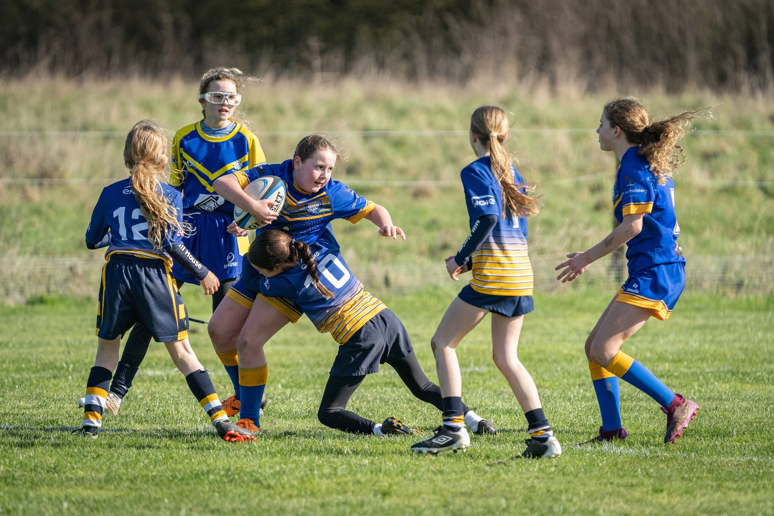 Young girls playing rugby on a grassy field, wearing blue and yellow uniforms, with one girl holding a rugby ball and others engaging in the game.