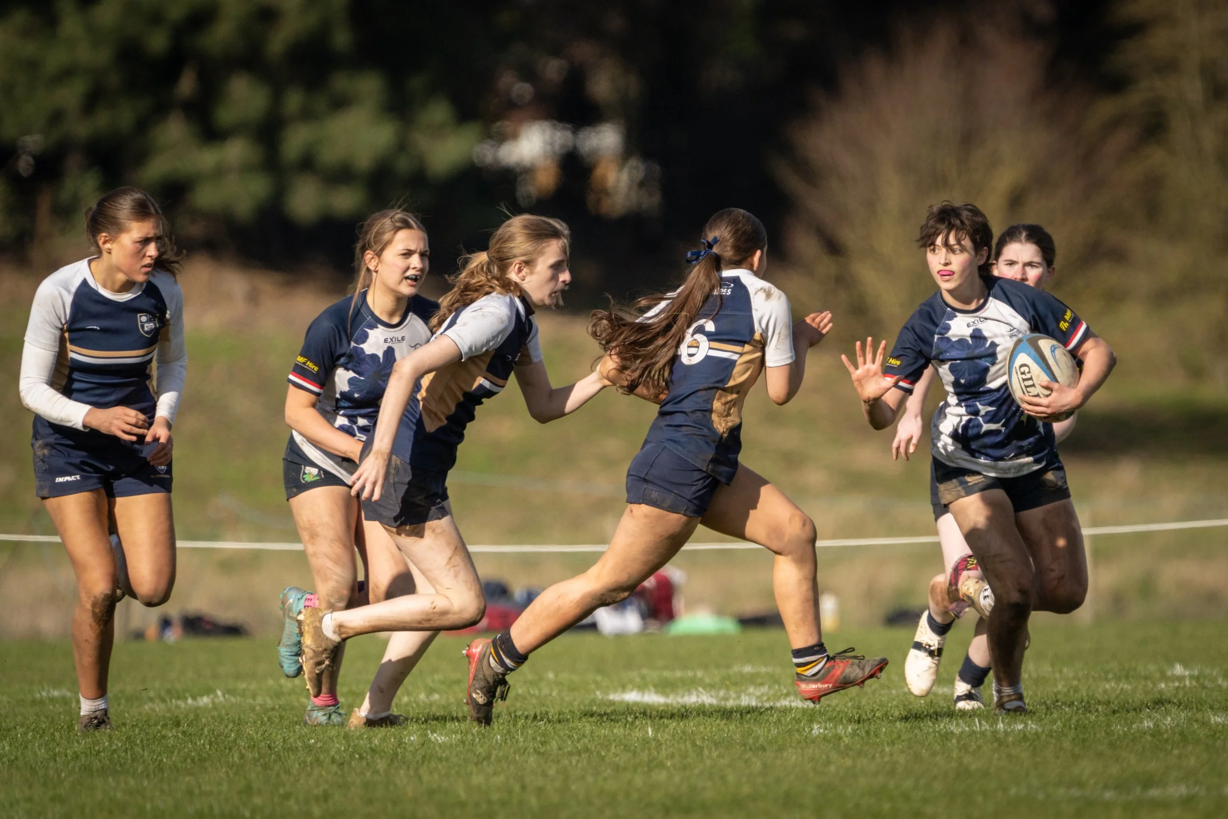 A group of young women playing rugby on a grassy field, some with mud on their legs, wearing blue and white sports uniforms, with one holding a rugby ball.