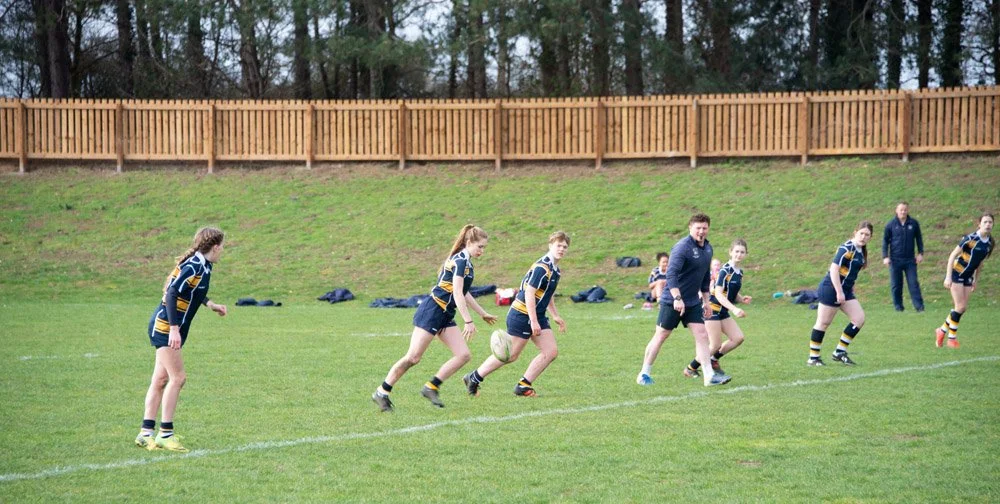 Women and girls in rugby uniforms practicing running on a grassy field during daytime.