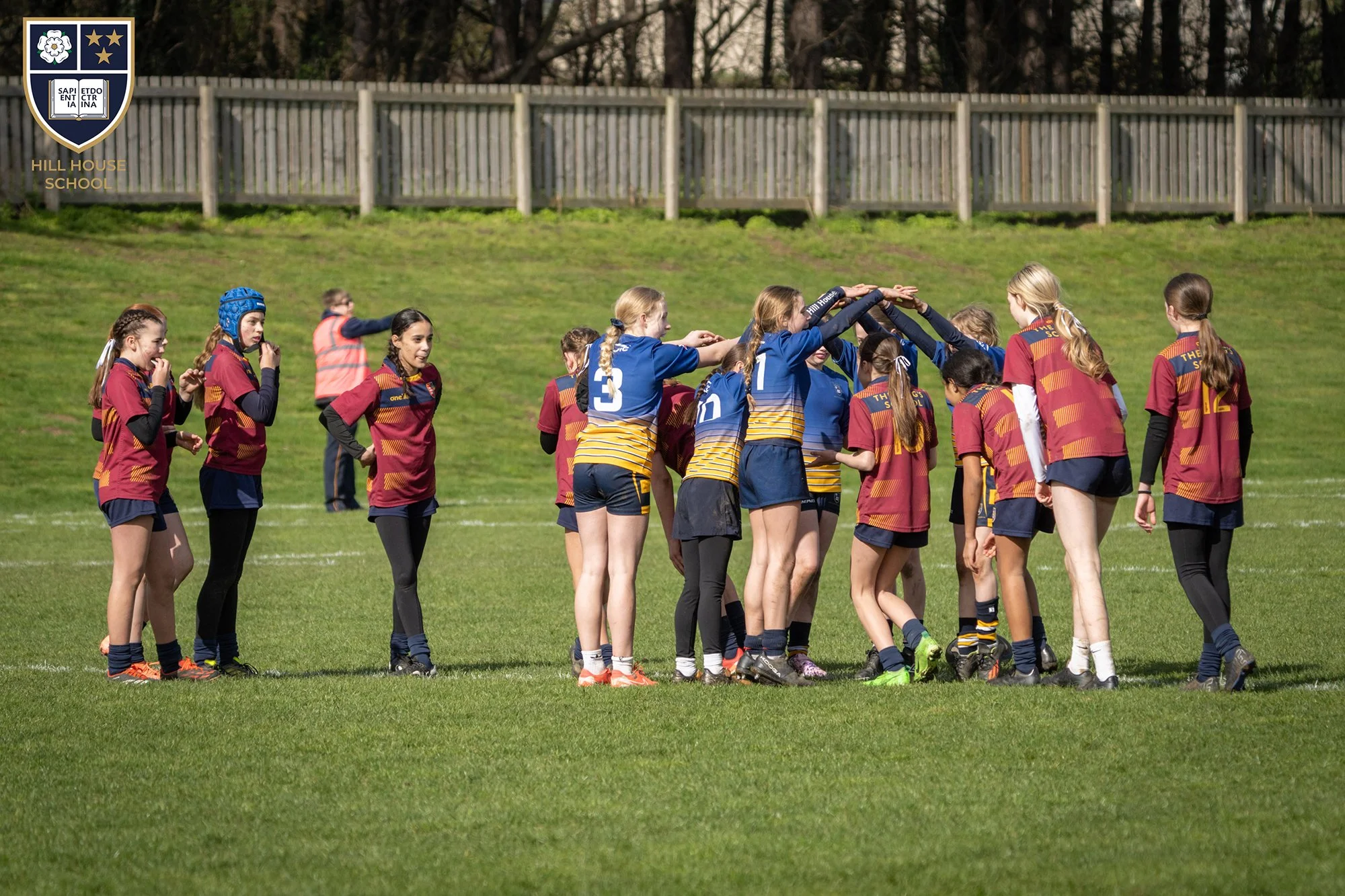 A group of female rugby players, wearing maroon and yellow uniforms, forming a tunnel with their arms for their teammate, who is wearing a blue and yellow uniform, to pass through during a game on a grassy field. A person in a pink vest is visible in