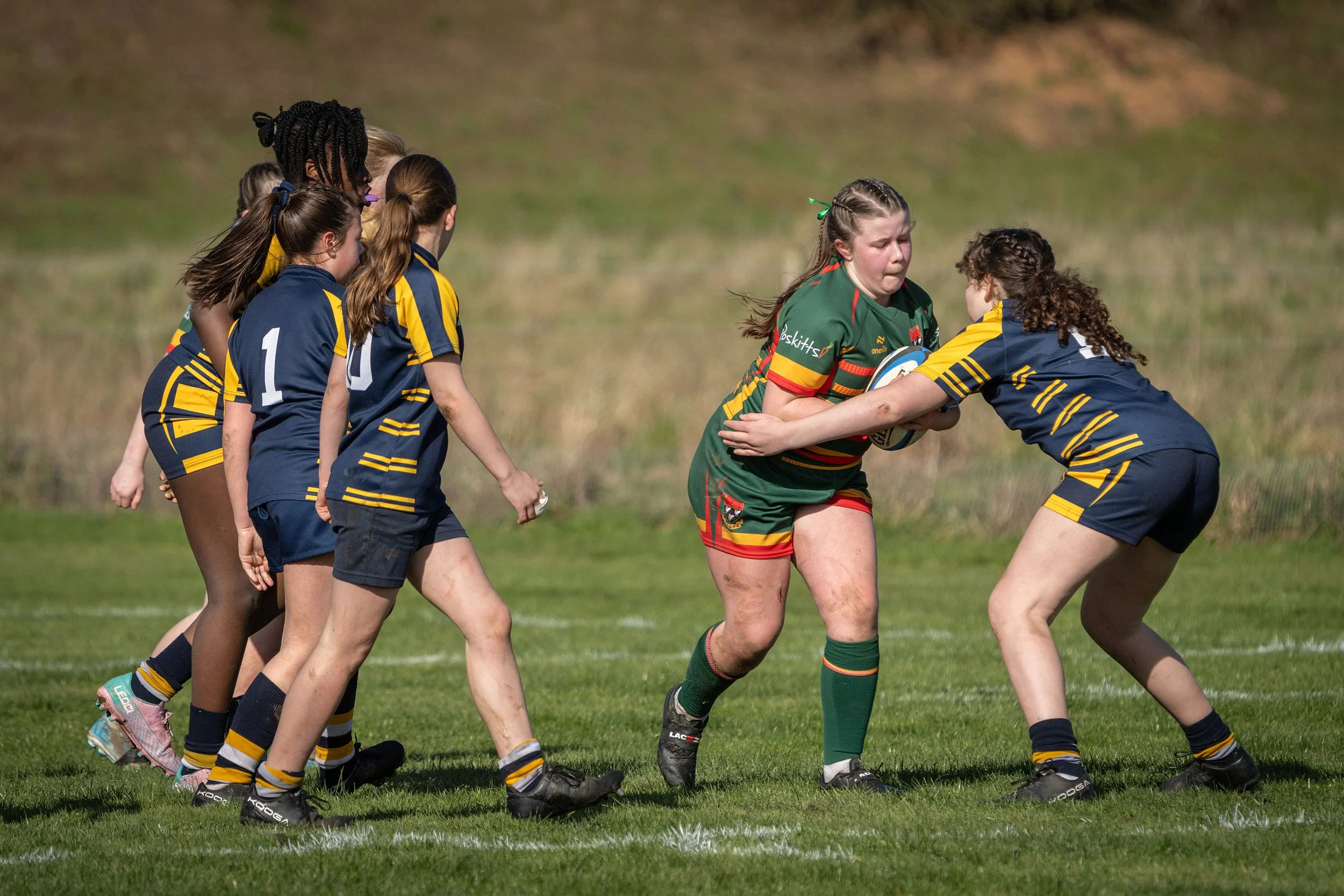 Women playing rugby on a grassy field, with some women in yellow and navy uniforms and others in green and red uniforms, engaging in a tackle during a game.