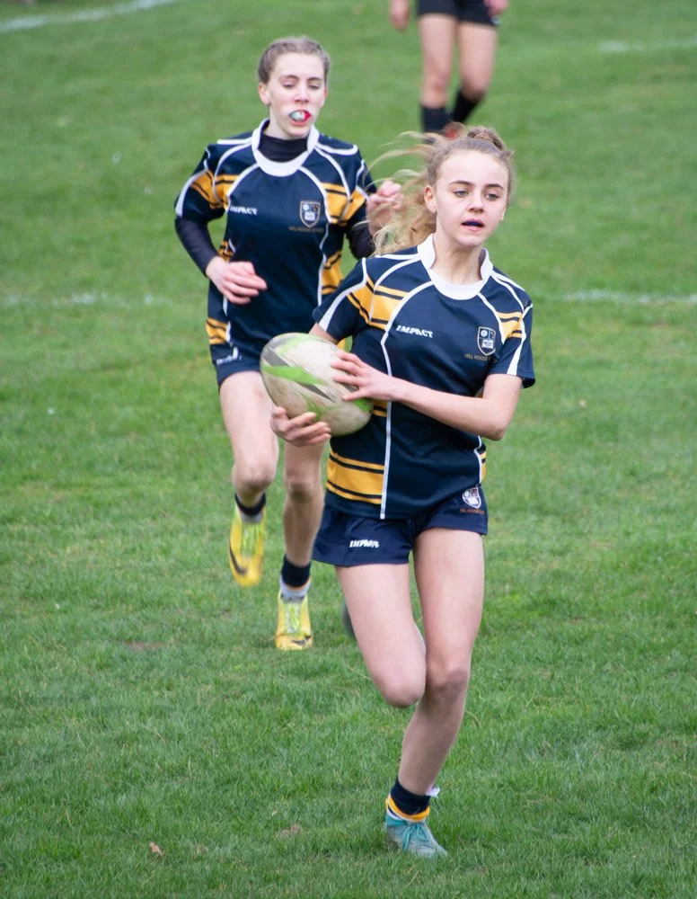 Two young female rugby players in navy and yellow uniforms running on a grassy field, with one holding a rugby ball.