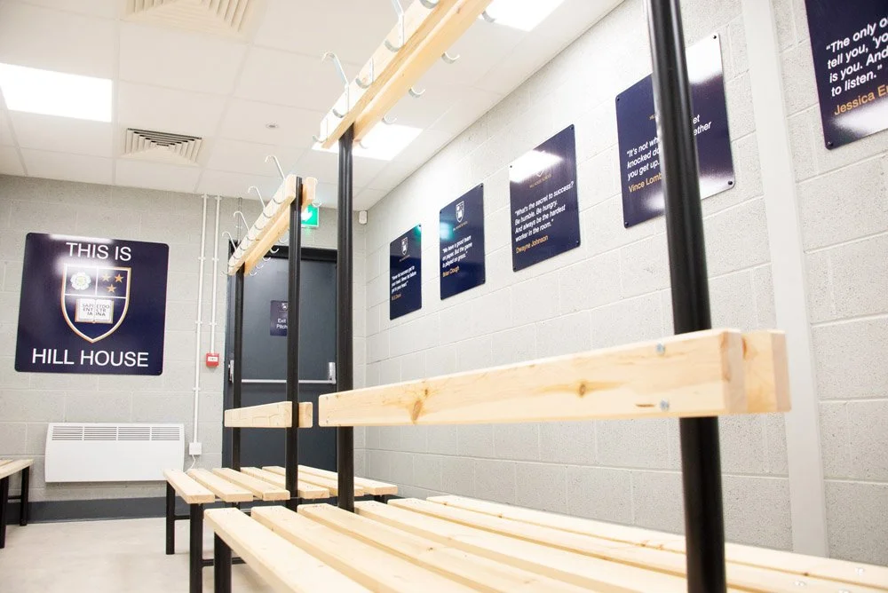 Empty wooden bench and coat rack inside a room with motivational quotes on the wall and a large sign that reads 'THIS IS HILL HOUSE' in a school or gym setting.