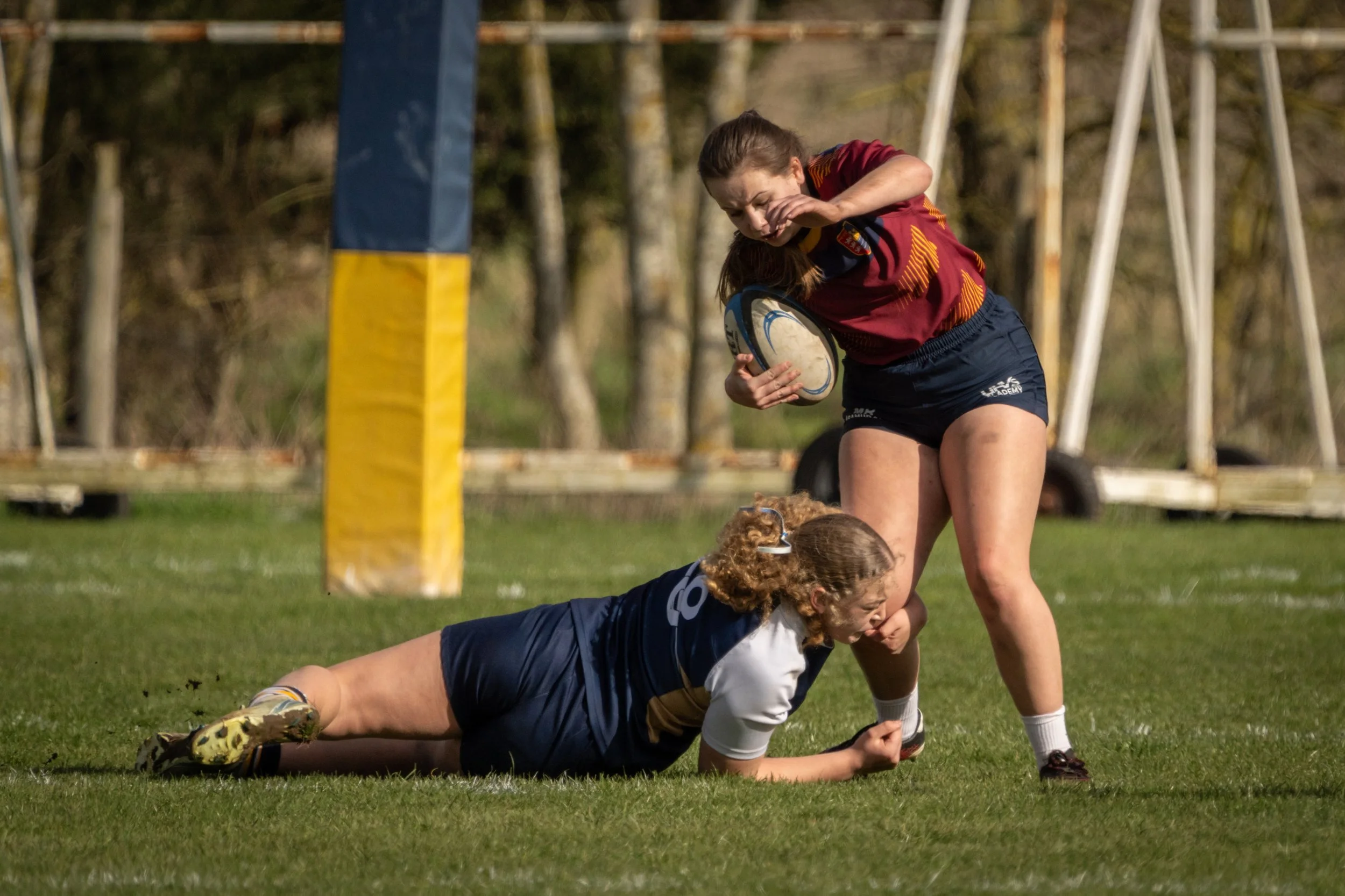 A rugby match with players in action on a grassy field, with one player tackled to the ground and another approaching.