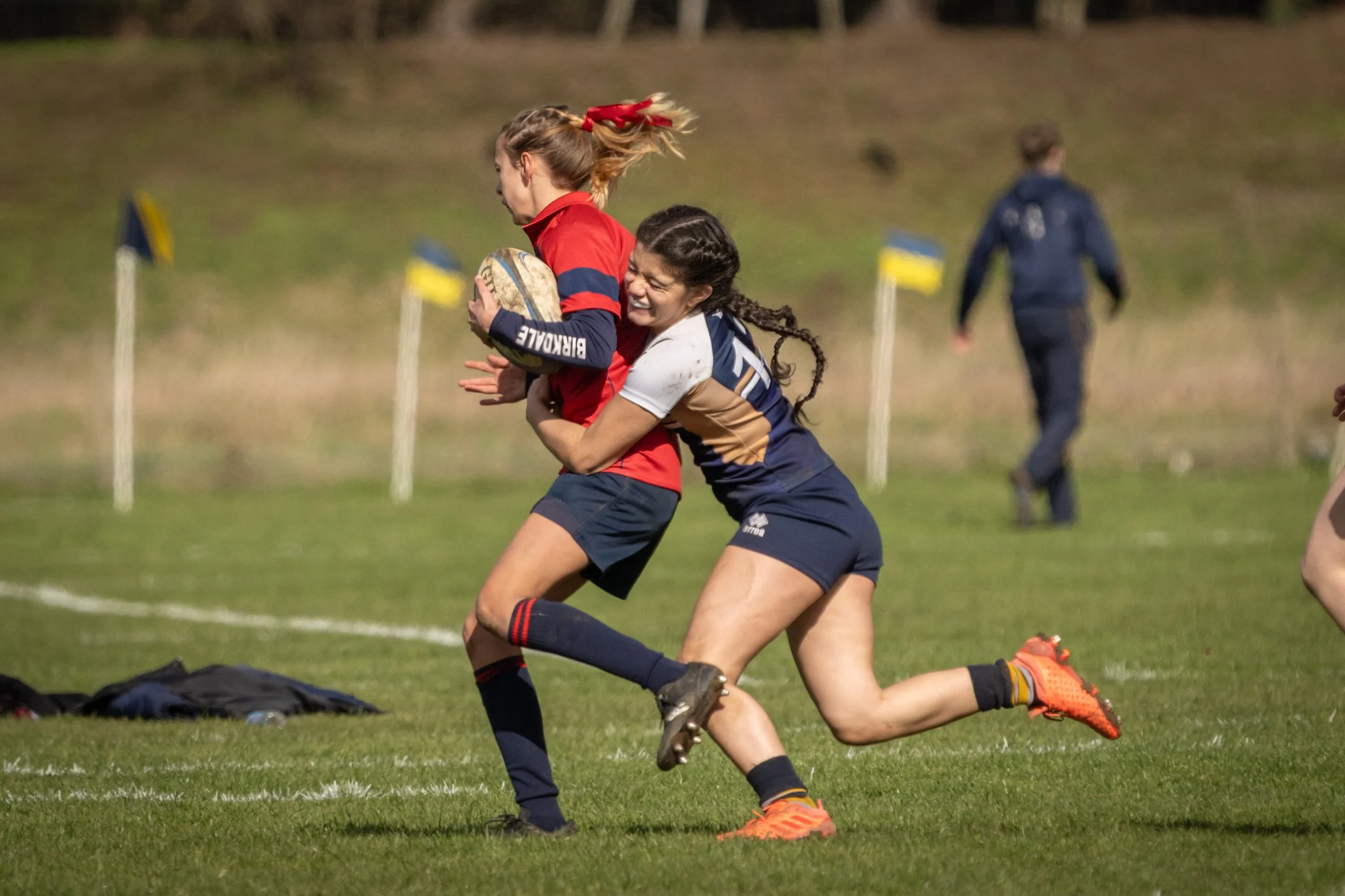 Two female rugby players are engaged in a tackle during a game on a grassy field, with one player holding a rugby ball. One player wears a red jersey and the other a navy and beige jersey, both focused and in motion.