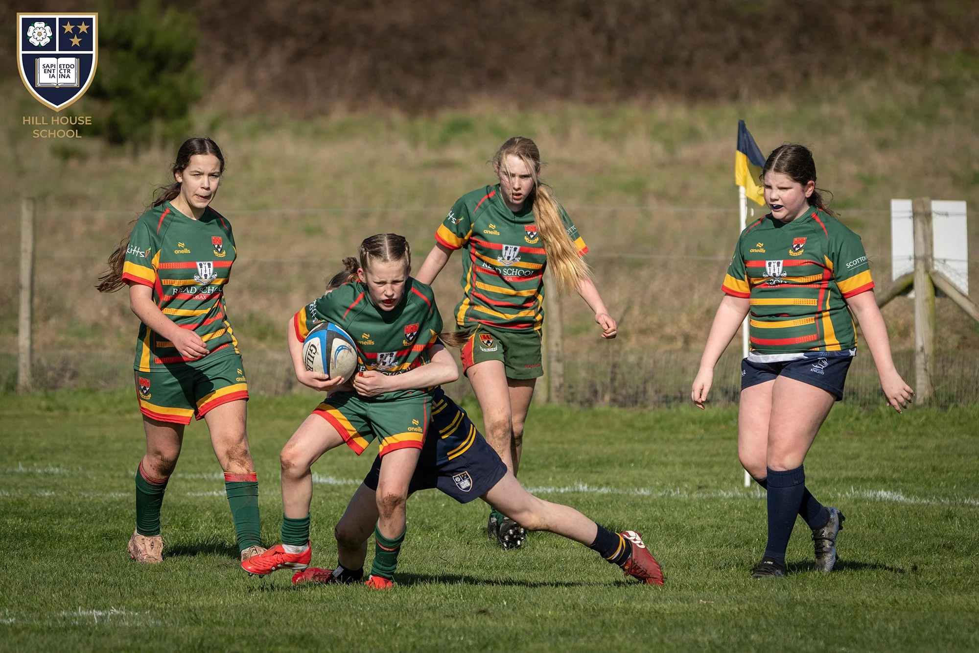 Girls playing rugby on a grassy outdoor field, wearing green uniforms with yellow and red accents, and one girl holding a rugby ball, with a fence and a flag in the background.