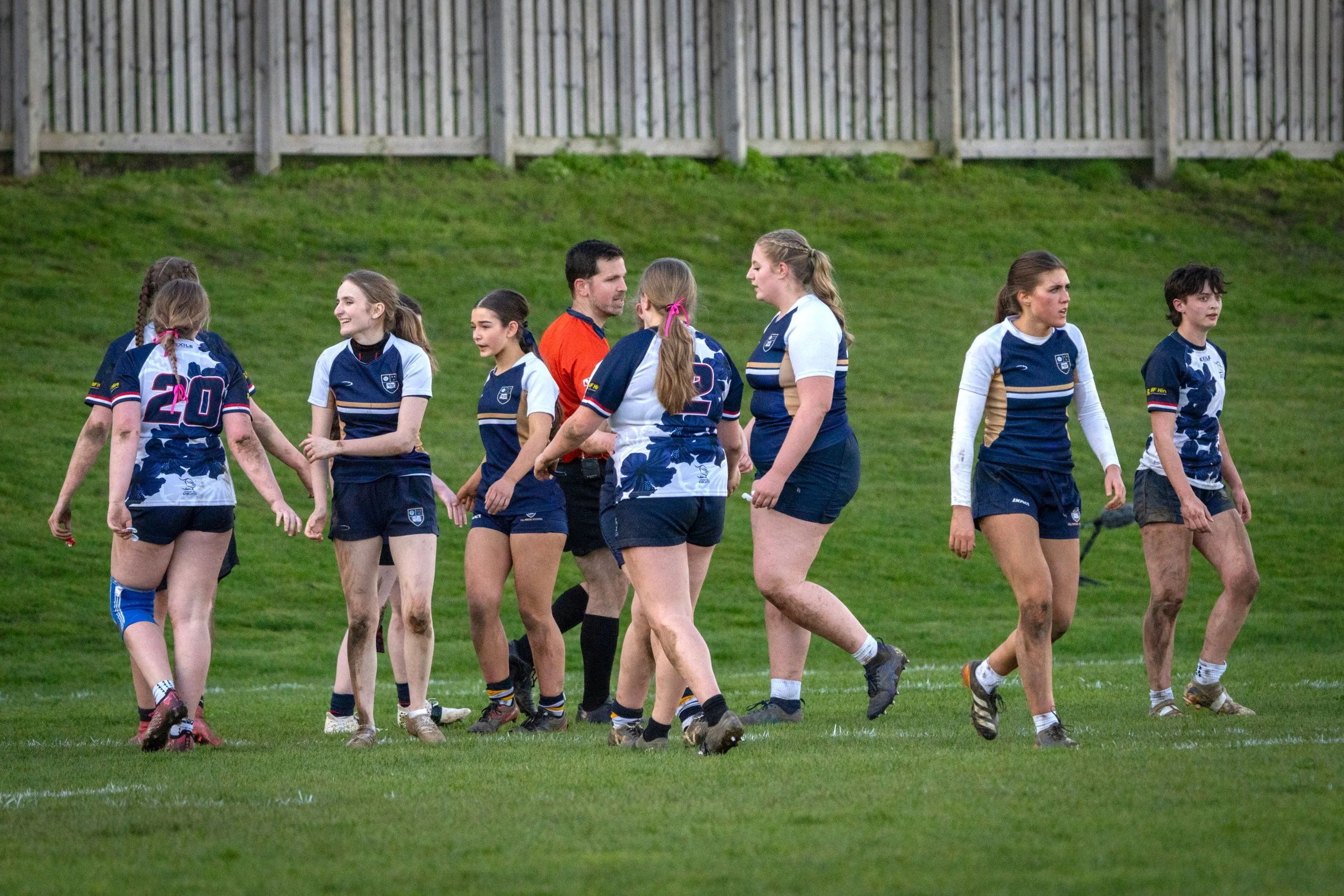 A group of young female rugby players and their coach on a grassy field, some with muddy legs, wearing blue and white uniforms, engaging in conversation after a game.
