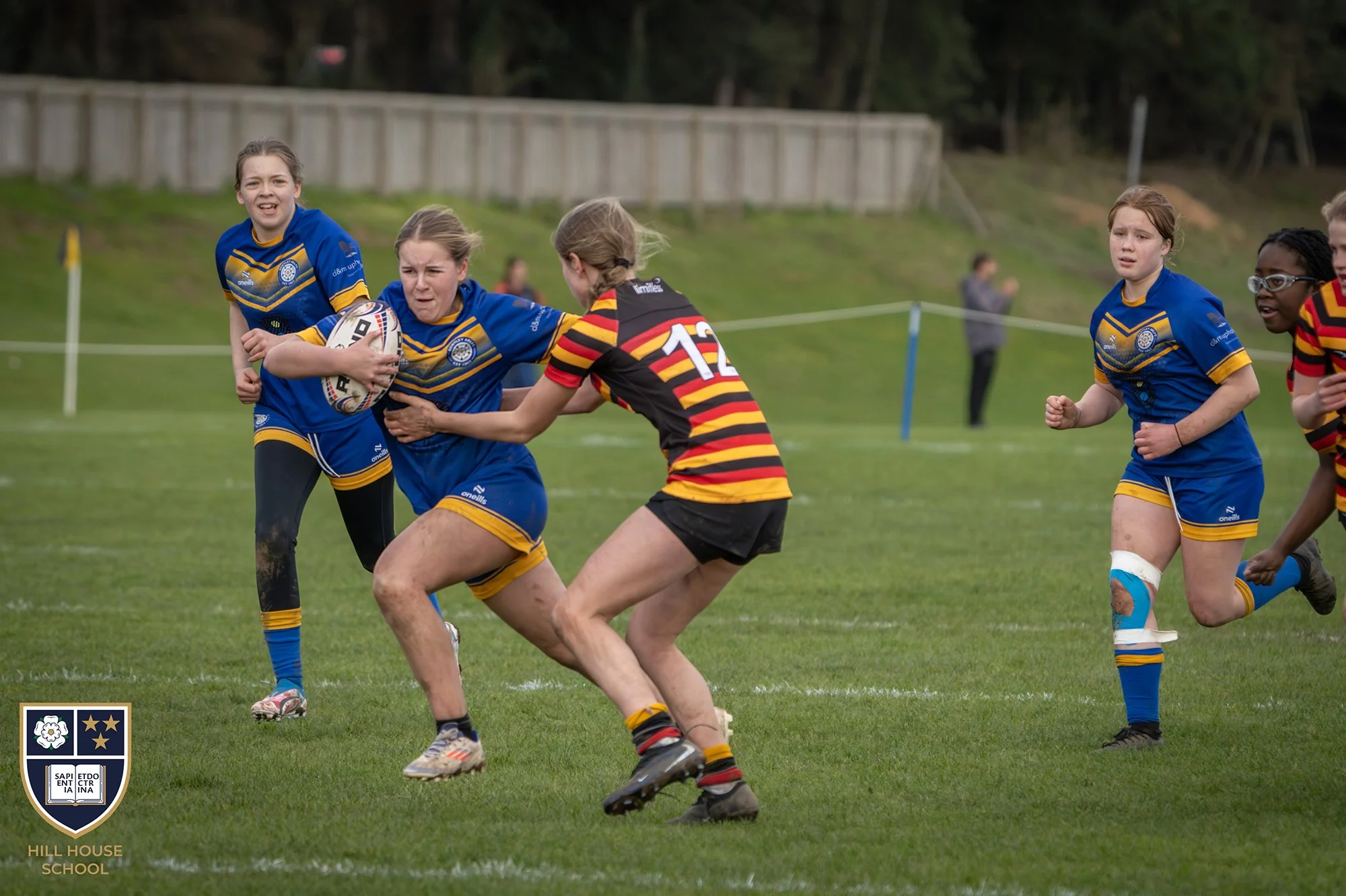 Girls playing rugby on a field, wearing blue and red/black striped uniforms. One girl in blue holds the rugby ball while being tackled by a girl in striped uniform. Other girls are running nearby. Fenced area and grassy hill in background.