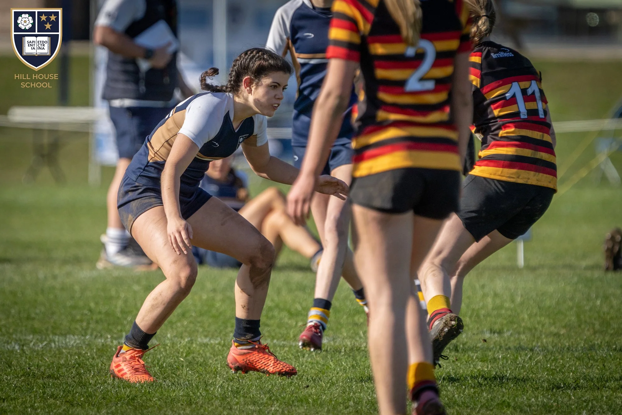 Young female rugby player crouching and preparing to tackle during a rugby match, with other players in striped uniforms and a grassy field in the background.