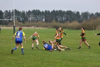 Rugby players in action on a grassy field, with some players tackling and others running, under a cloudy sky.