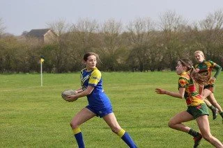 Girls playing rugby on a grassy field, with one girl in yellow and blue holding the ball and others in colorful jerseys running after her.