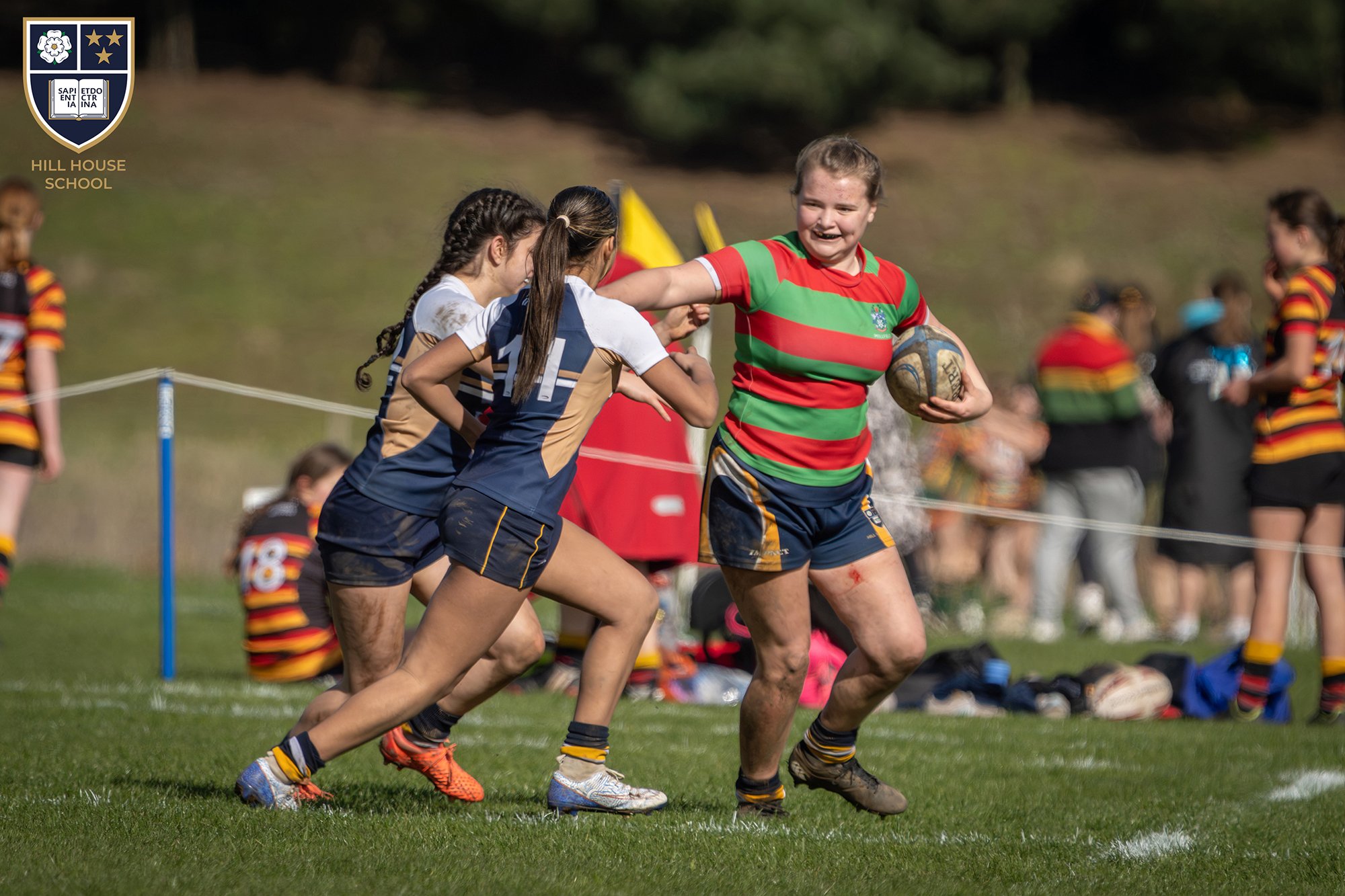 Young female rugby players playing a match on a grassy field, with one girl in a red and green striped jersey holding a rugby ball, and other players in blue and tan jerseys attempting to block her, with spectators in the background.