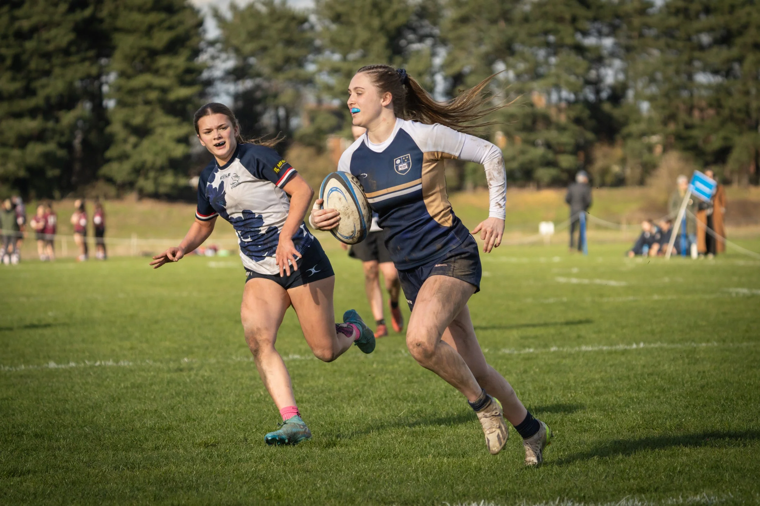 Two female rugby players running on a grassy field during a game, with trees in the background.