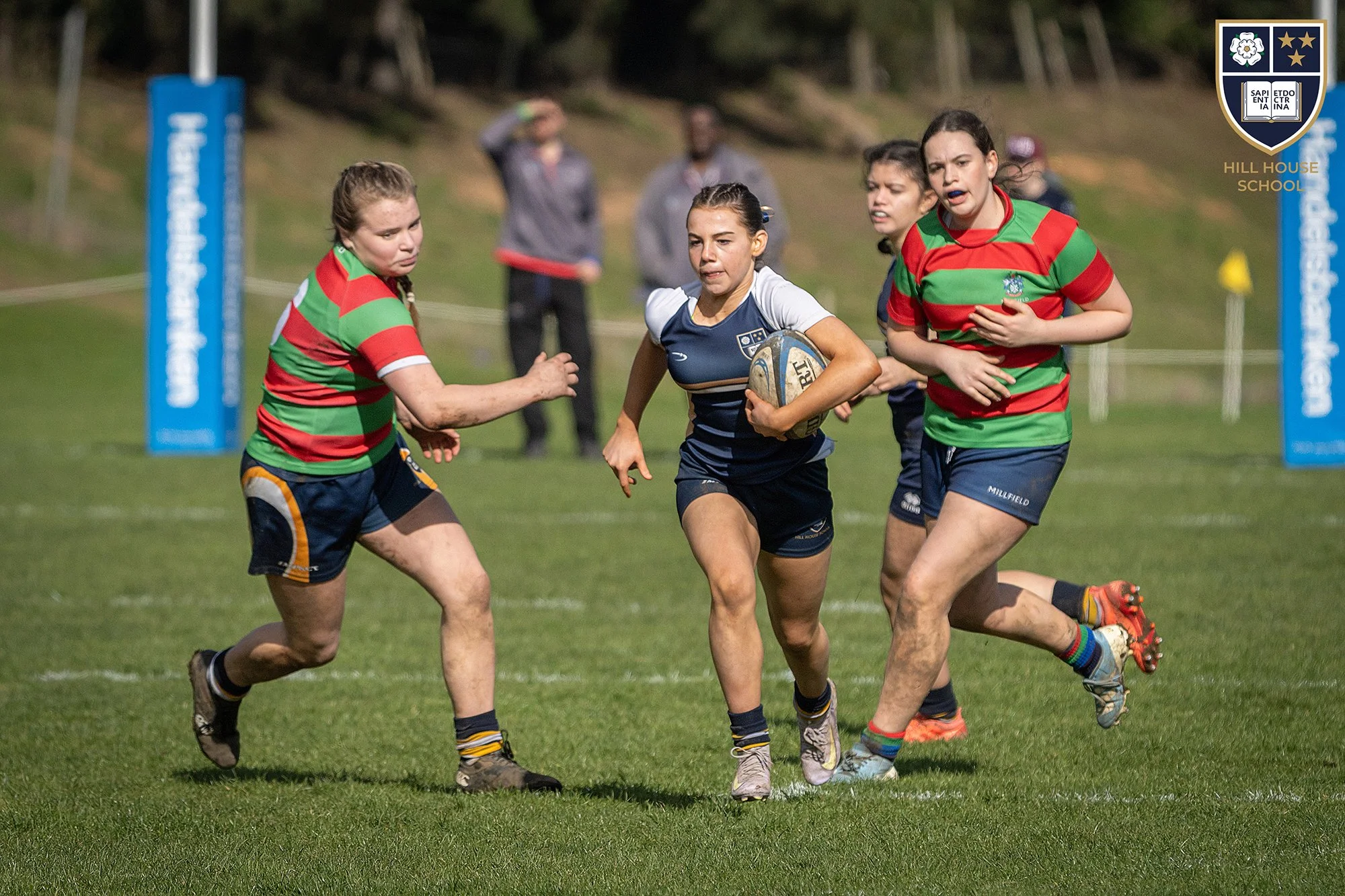 Girls playing rugby on a field, with three of them wearing red and green striped jerseys and one in a blue and white jersey, running with a rugby ball. In the background, there are two people watching and a school emblem in the top right corner.