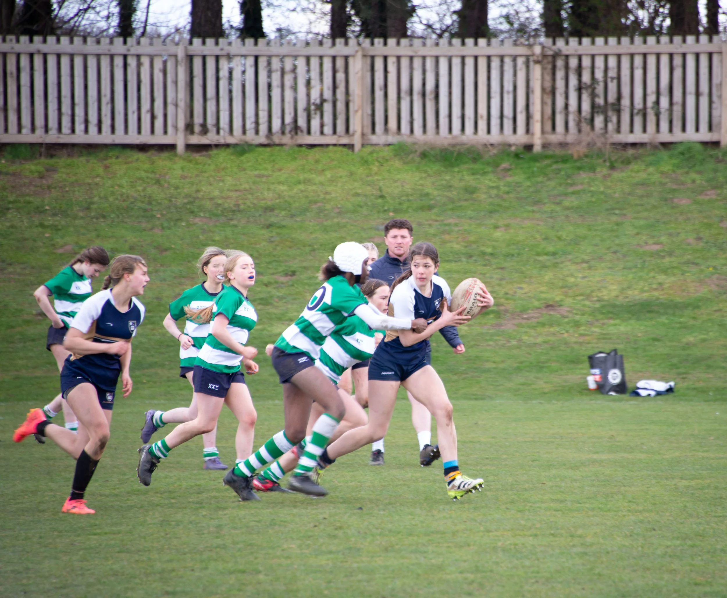 A group of young women playing rugby on a grassy field, with a wooden fence and trees in the background.