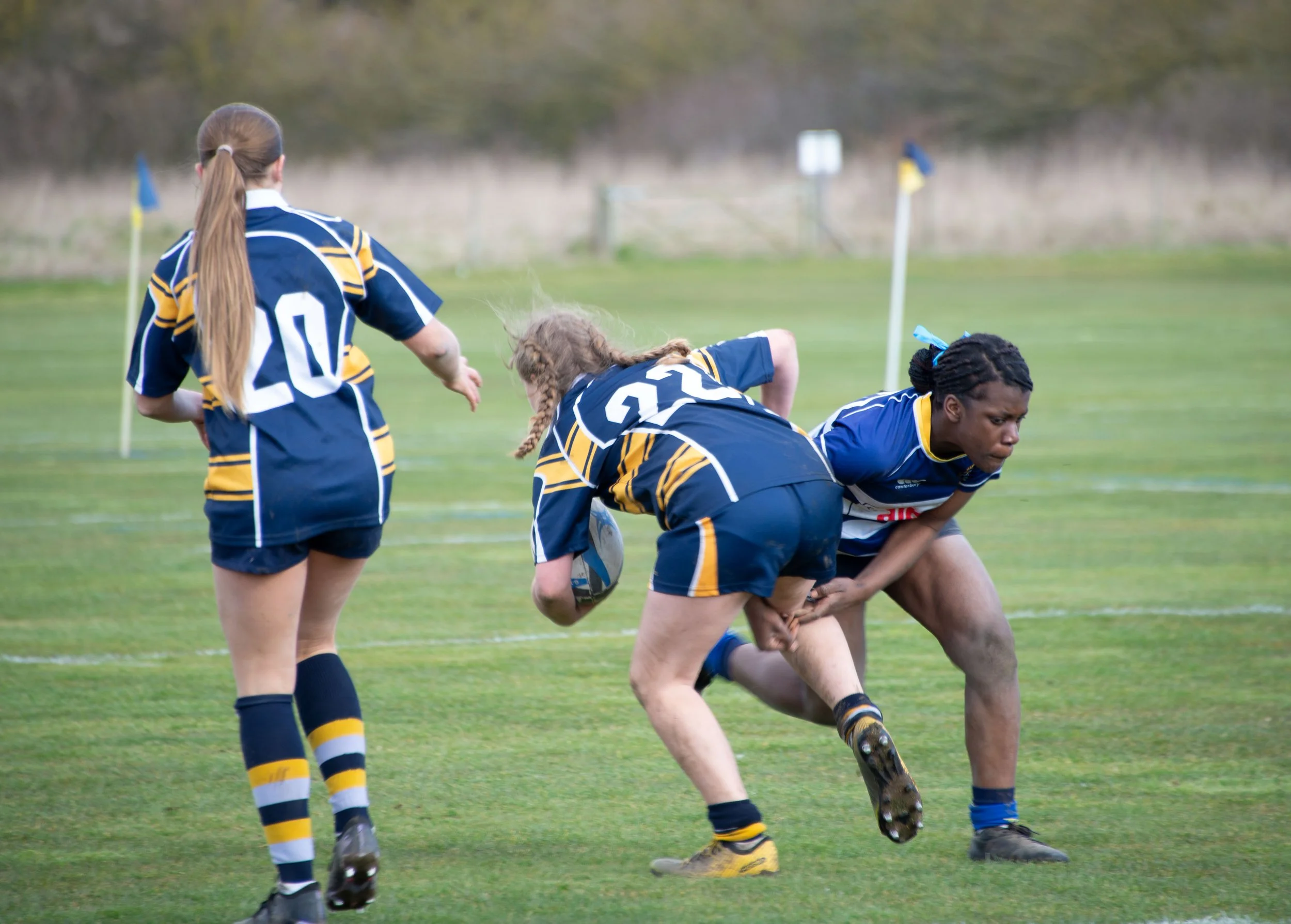 Women in sports uniforms playing rugby on a grass field, with some women engaged in a scrum and others observing.
