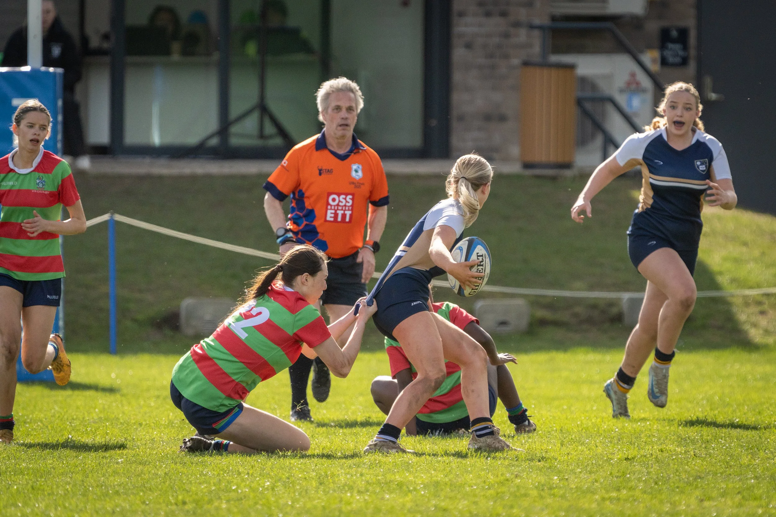 Women playing rugby on a grassy field, some women are kneeling or running, and one woman is holding a rugby ball. A man in an orange jersey watches nearby.