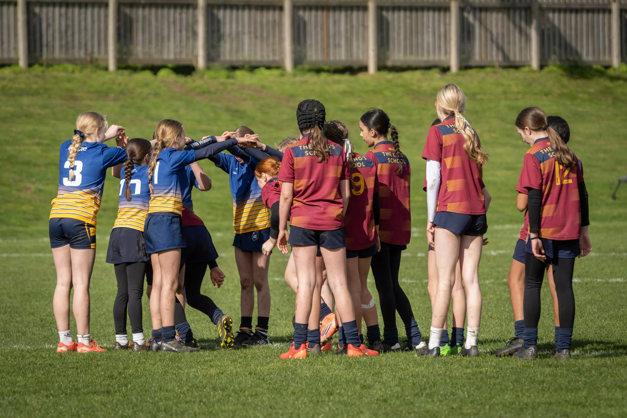 Girls' sports team huddled together on a green field, wearing blue and maroon jerseys with numbers, during a game or practice.