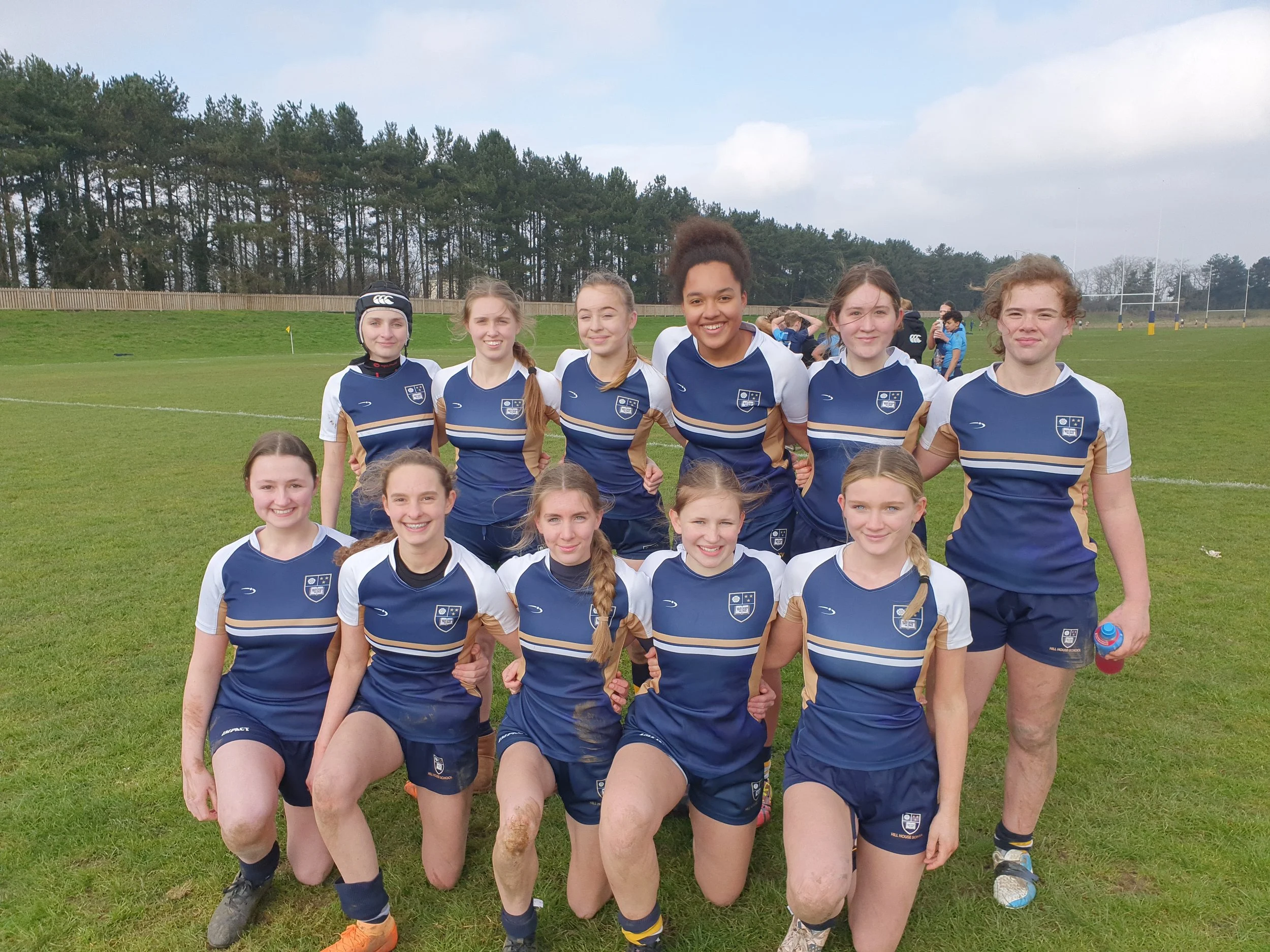 A group of young female rugby players wearing blue and white uniforms with a crest, posing outdoors on a grassy field with a wooden fence and trees in the background.