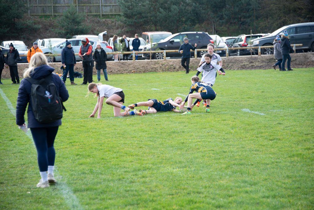 Children playing rugby on a grassy field, with spectators watching from the sidelines and parked cars in the background.