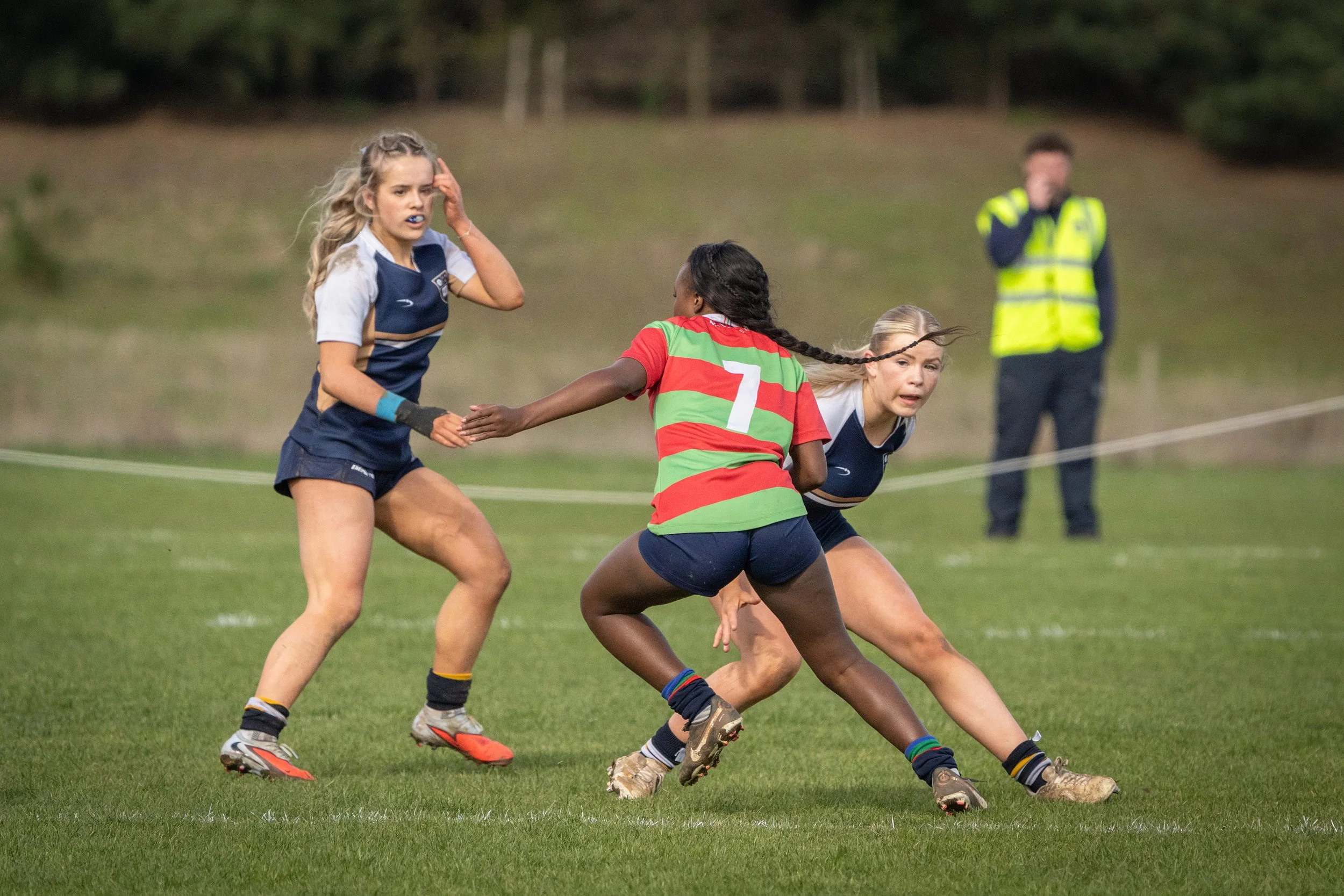 Three young female rugby players in action on a grassy field, dressed in sports gear including jerseys, shorts, and cleats, with a referee or coach in a bright yellow vest in the background.