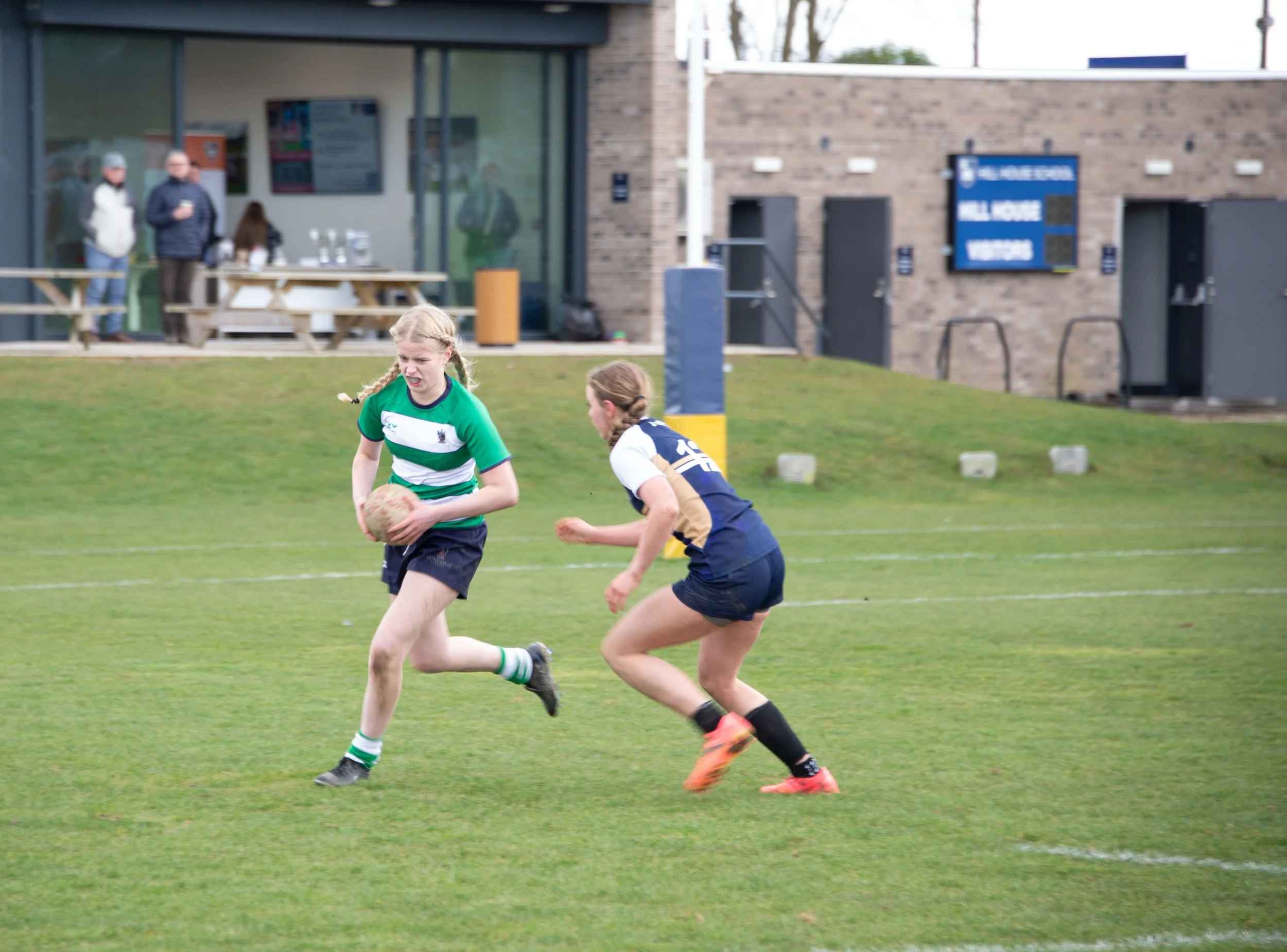 Two young girls playing rugby on a grassy field with a building and a sign in the background. One girl, wearing a green and white jersey, is holding the rugby ball, while the other, in a dark jersey with white sleeves, is running alongside her. In th